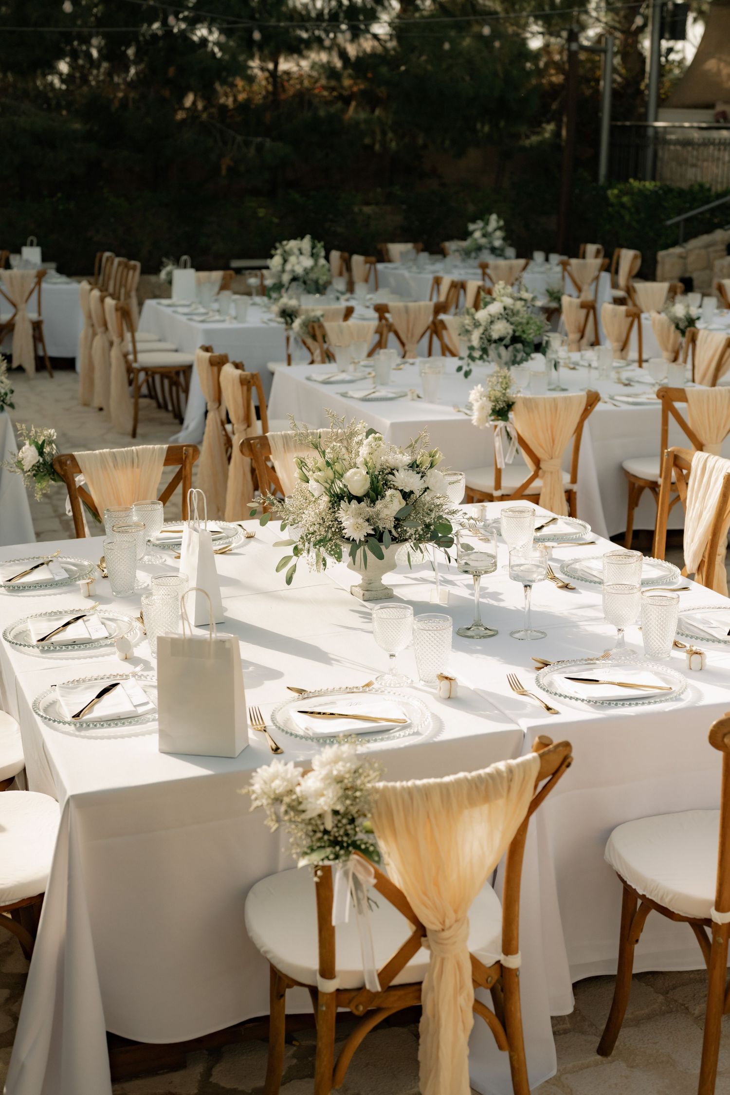 Elegant outdoor wedding reception with white tablecloths, wooden cross-back chairs, and white floral centerpieces.
