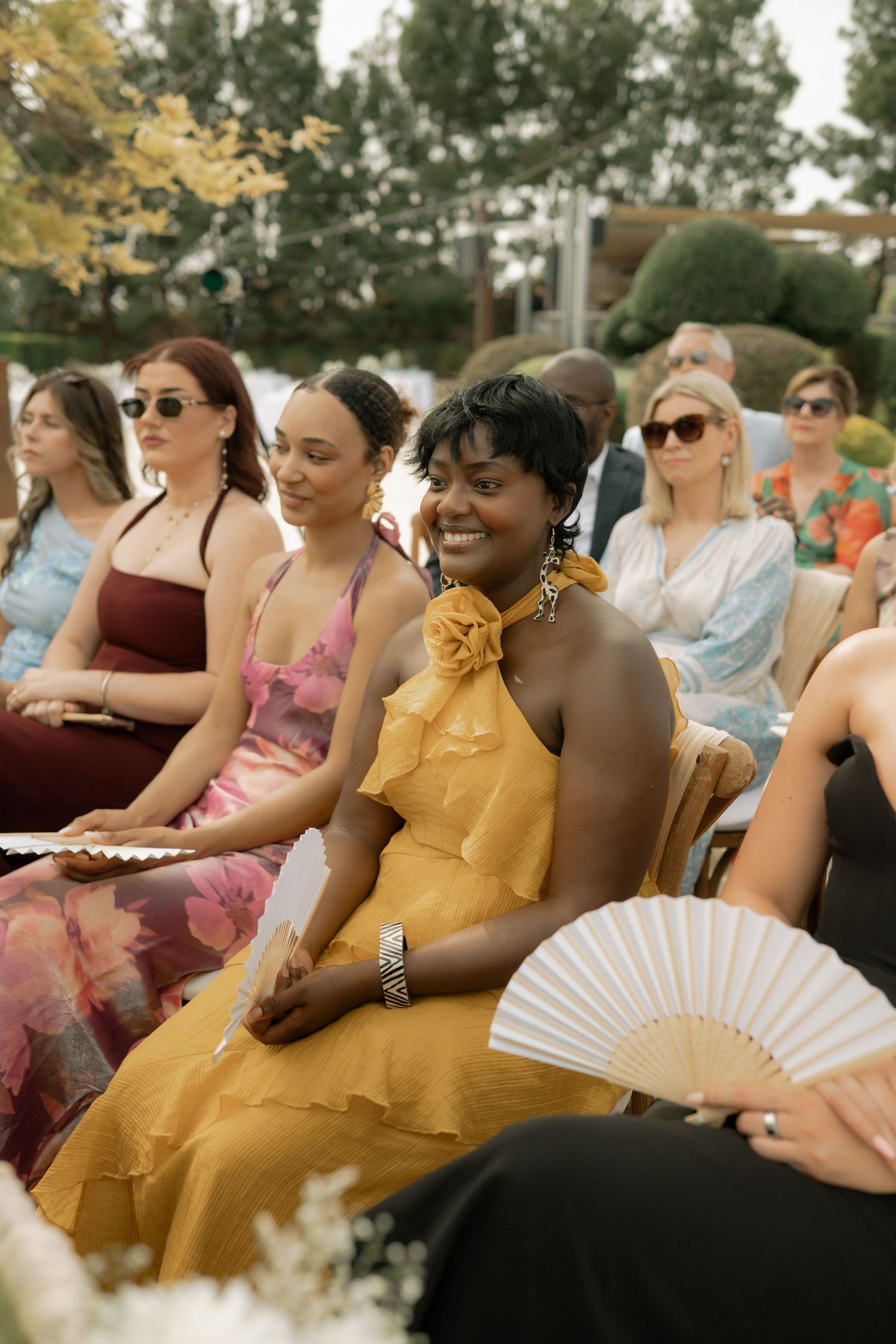 Woman in yellow dress smiling at outdoor wedding ceremony, holding a white hand fan, surrounded by guests.