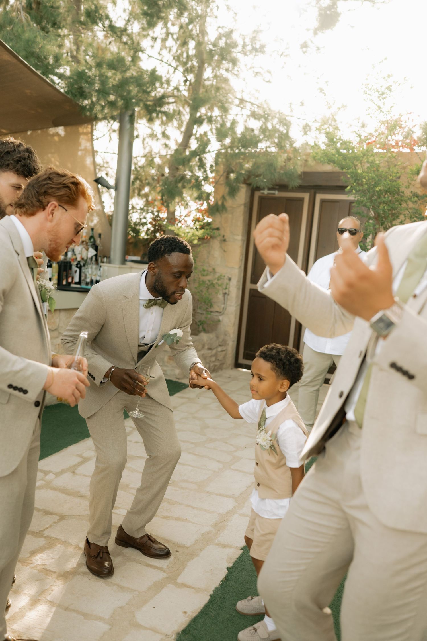 Groomsmen in tan suits dancing joyfully with a young boy at an outdoor wedding celebration.