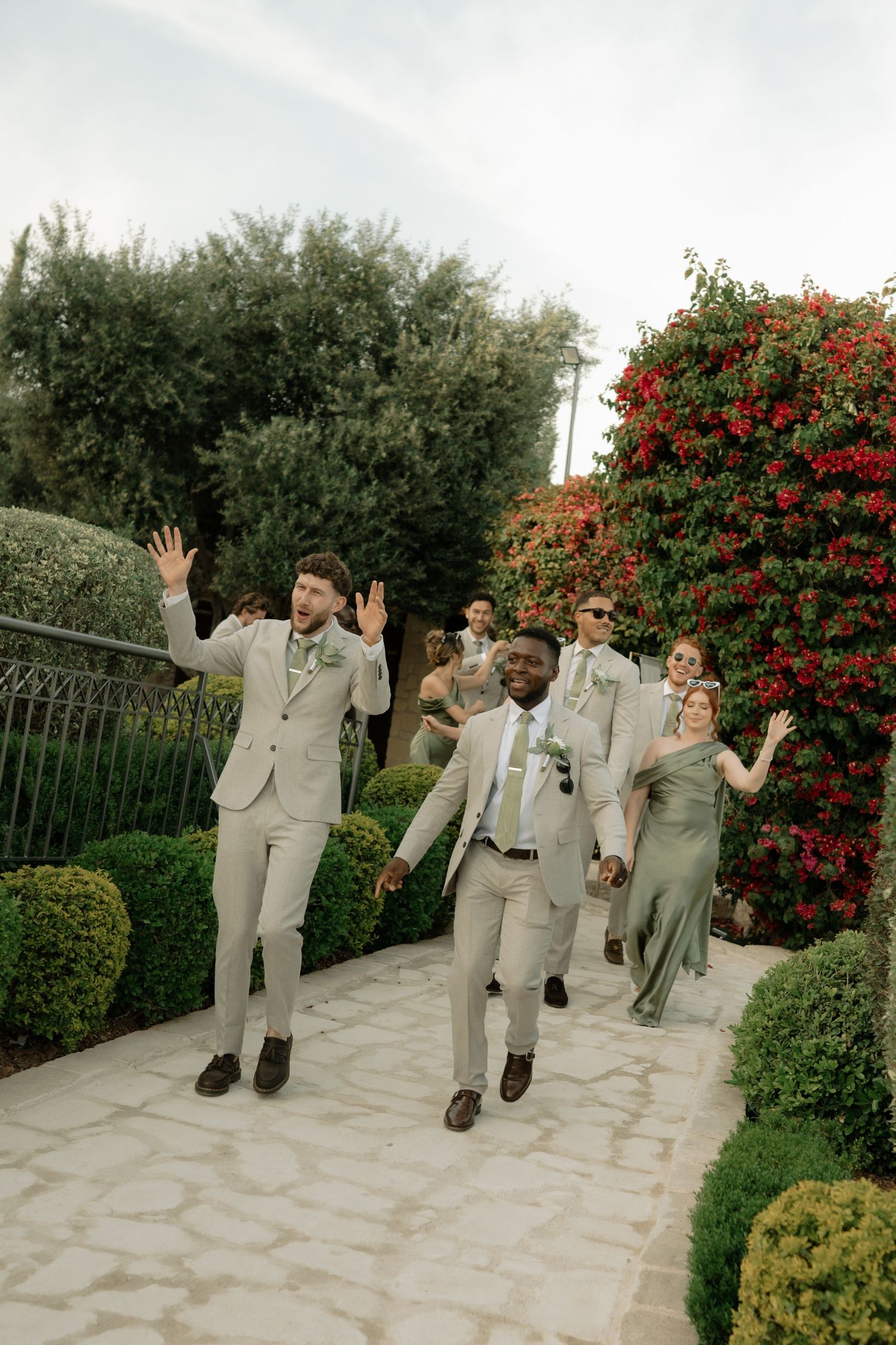 Groomsmen and bridesmaids in tan suits and sage dresses wave and celebrate outdoors at a garden wedding venue.