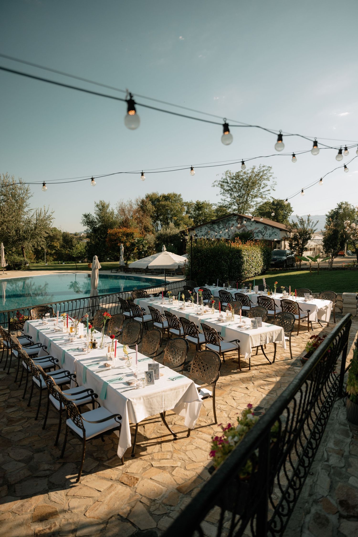 Elegant outdoor dining setup with string lights and long tables arranged on a stone patio overlooking a pool.