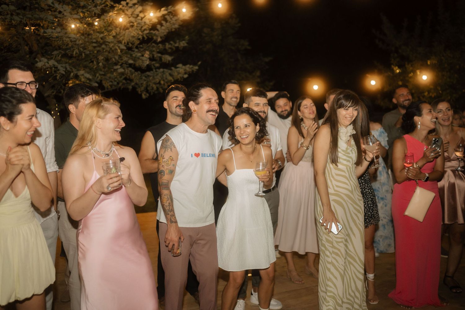 Wedding guests dance and celebrate together on an outdoor dance floor illuminated by string lights at night.