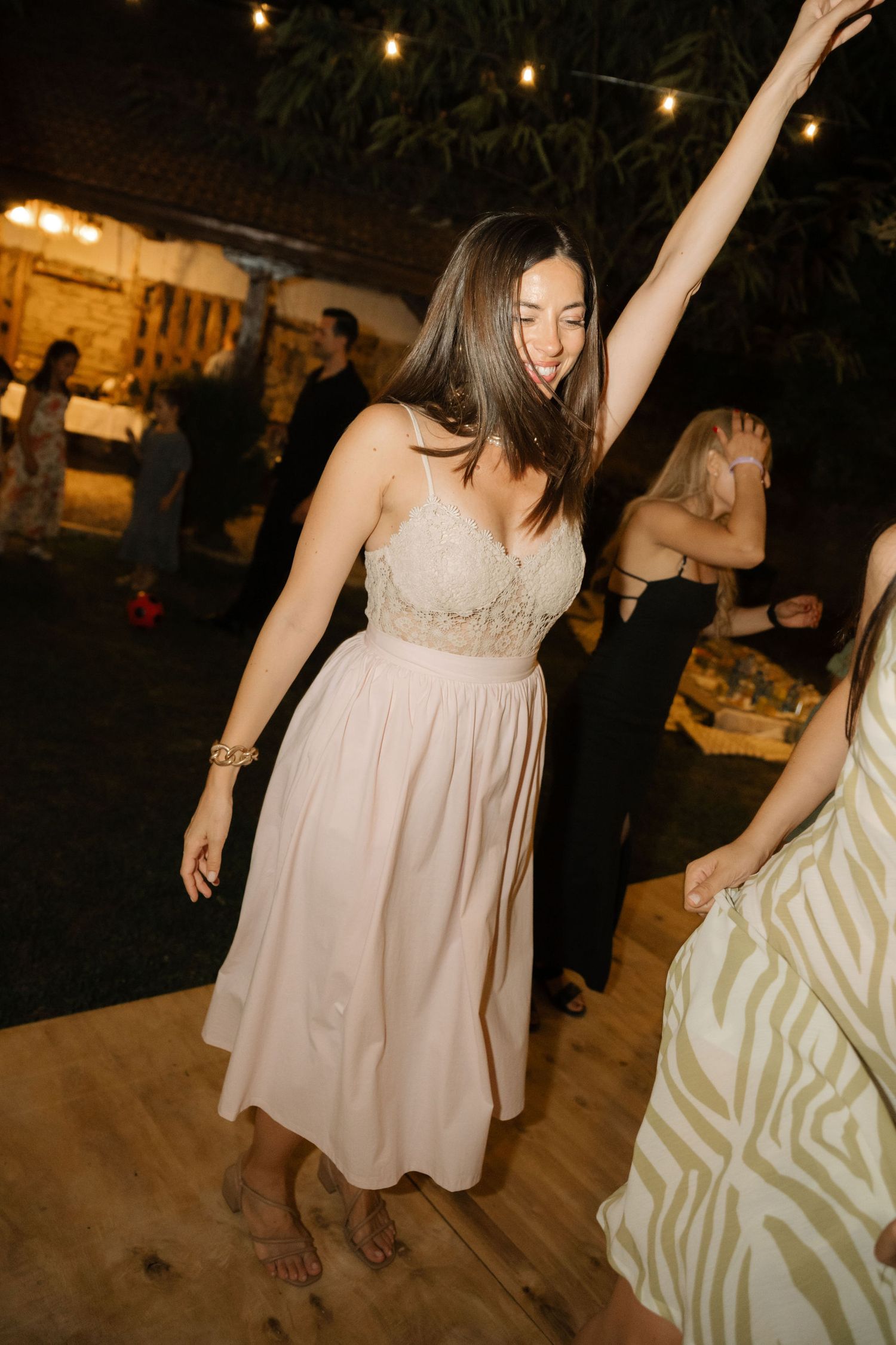 Person in a blush pink bridesmaid dress dances joyfully at an outdoor evening reception with string lights overhead.