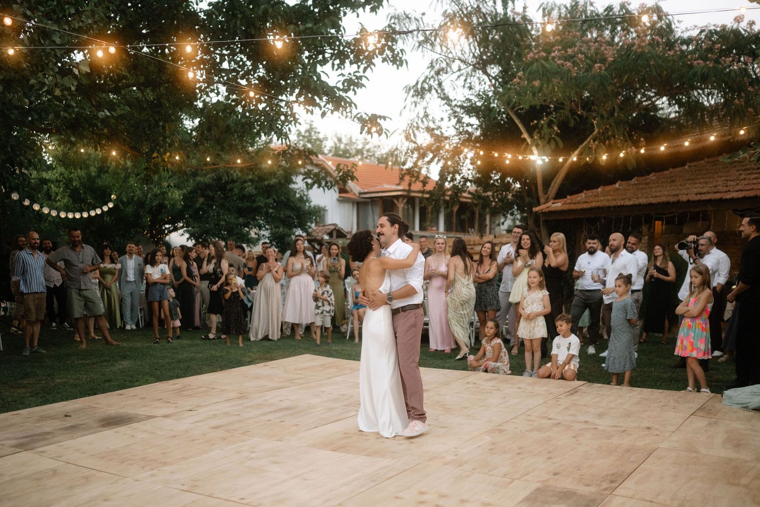 String lights illuminate an outdoor wedding reception area with guests gathered under a warm evening sky.