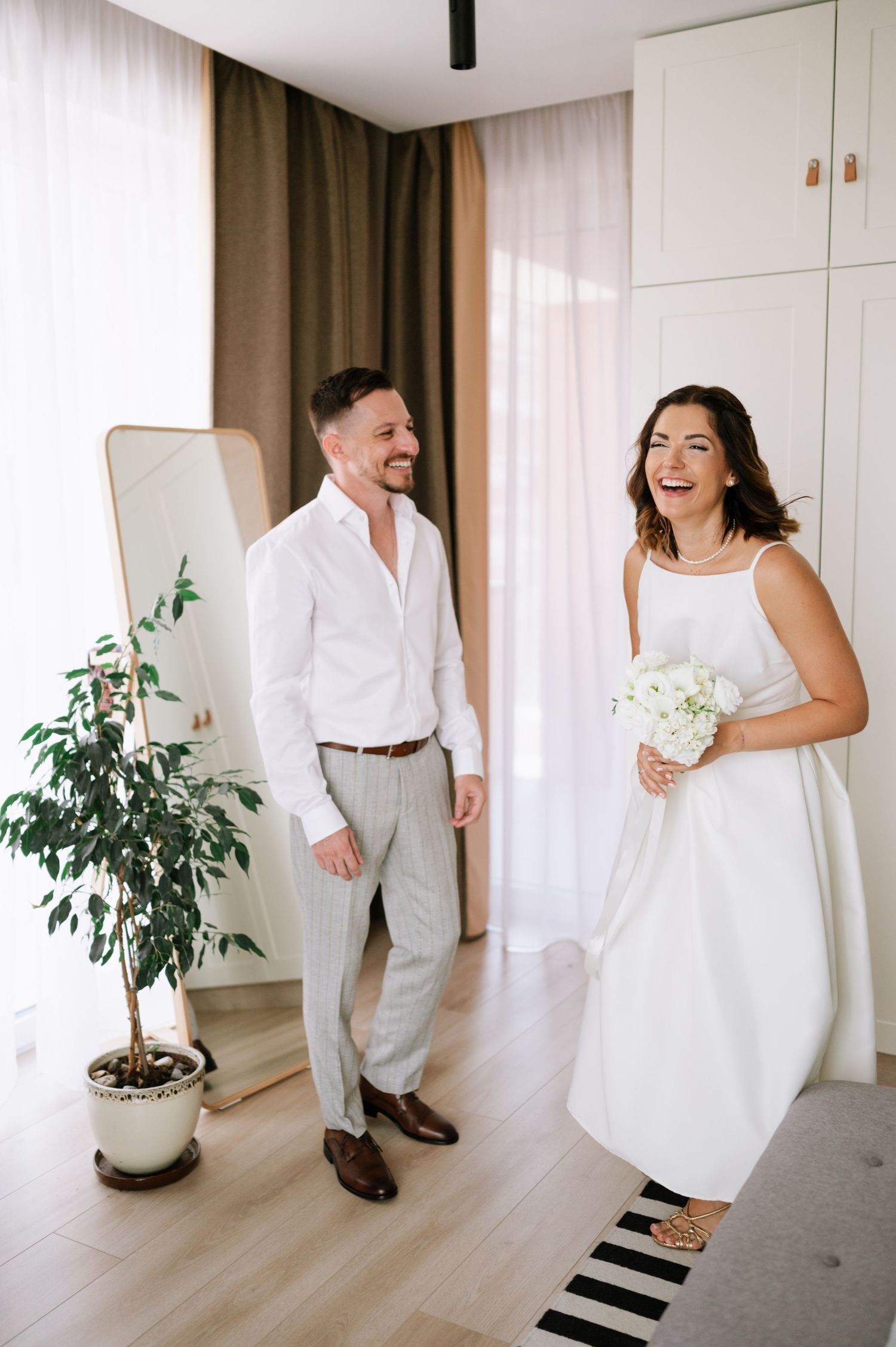 A joyful couple shares a candid moment of laughter in a bright modern room with white decor and a potted plant.
