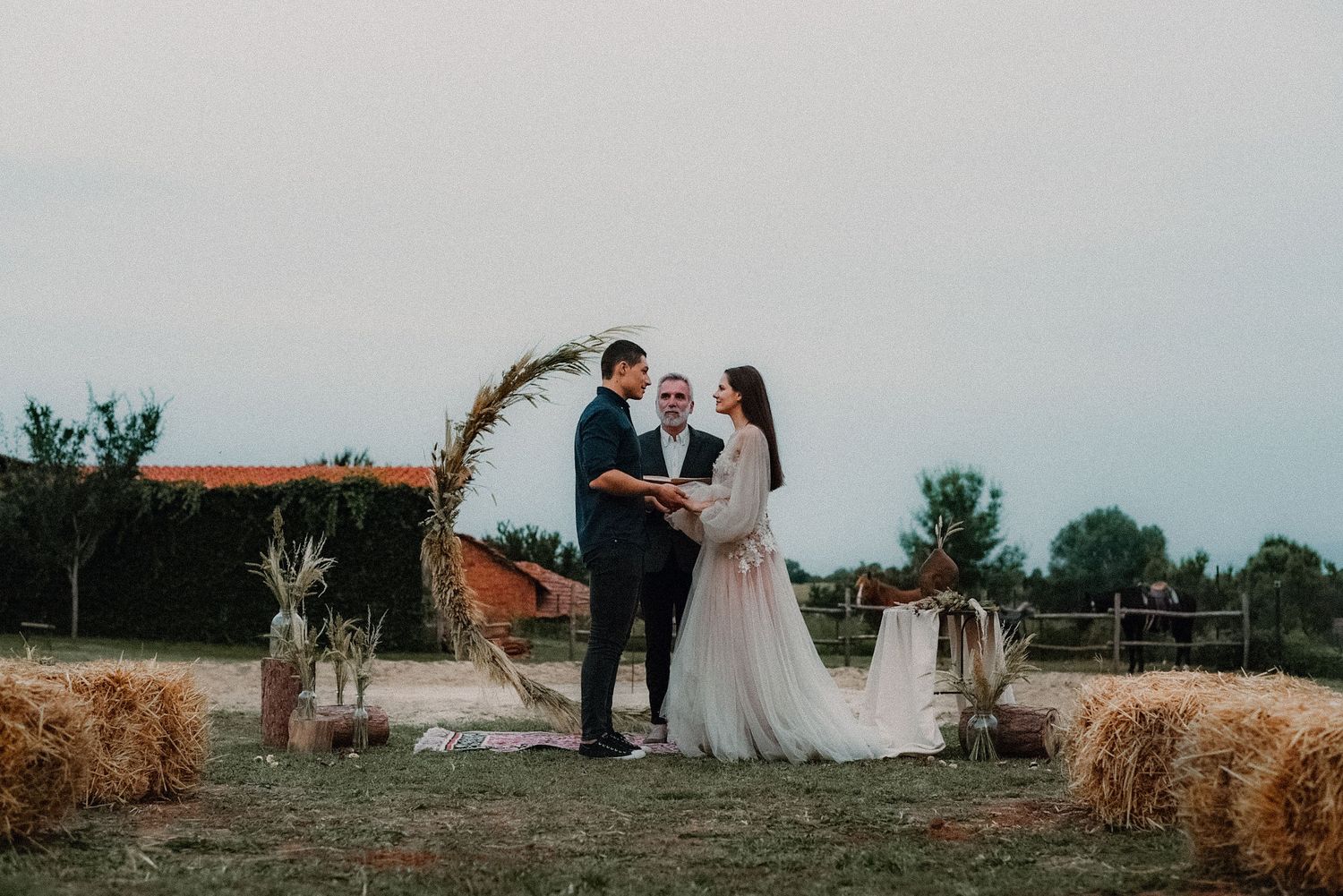 A couple shares a romantic moment at a rustic outdoor wedding ceremony with hay bales and an arch at sunset.