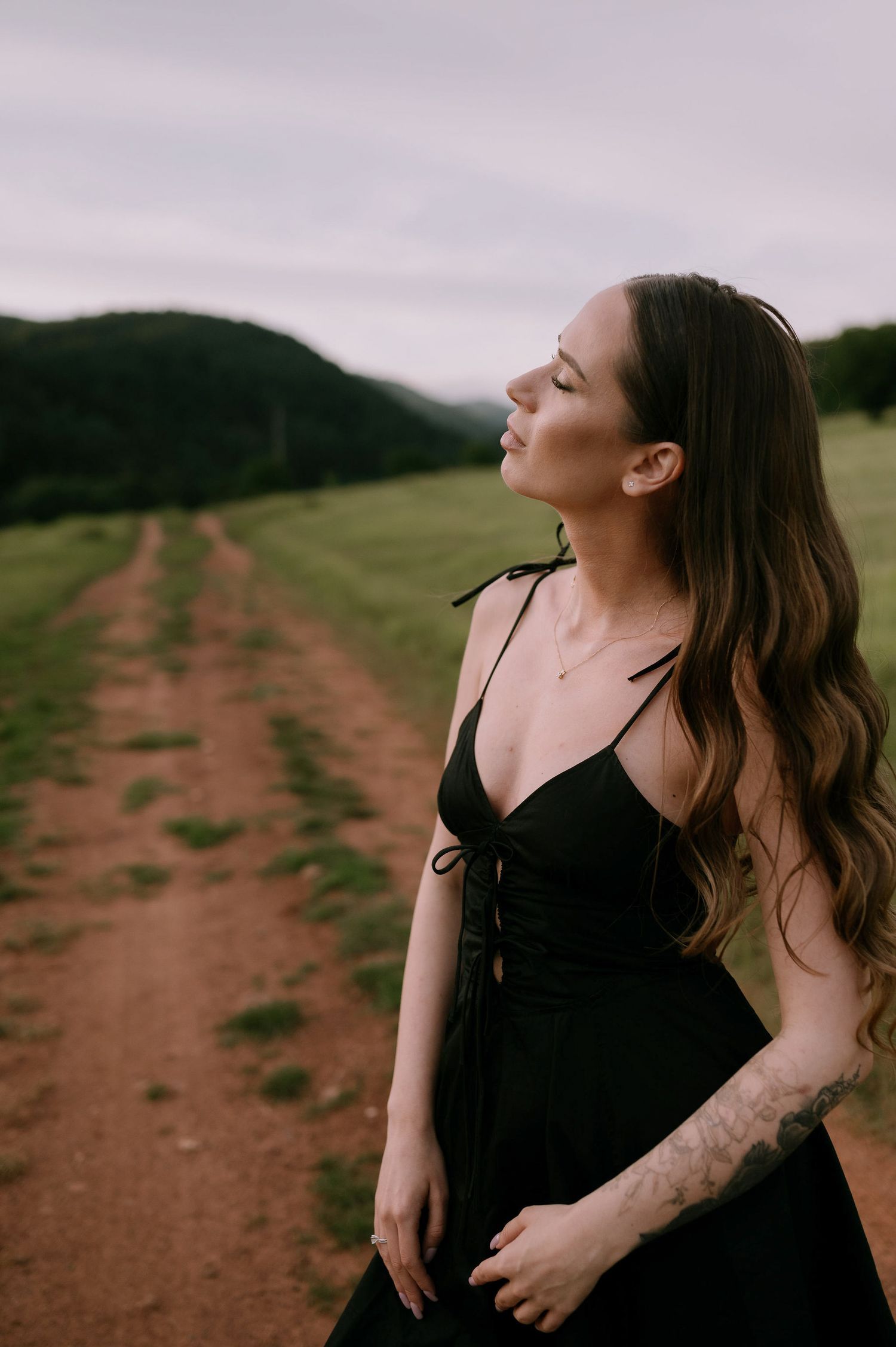 Woman in black dress laughs joyfully while standing in a rural field with mountains in background.