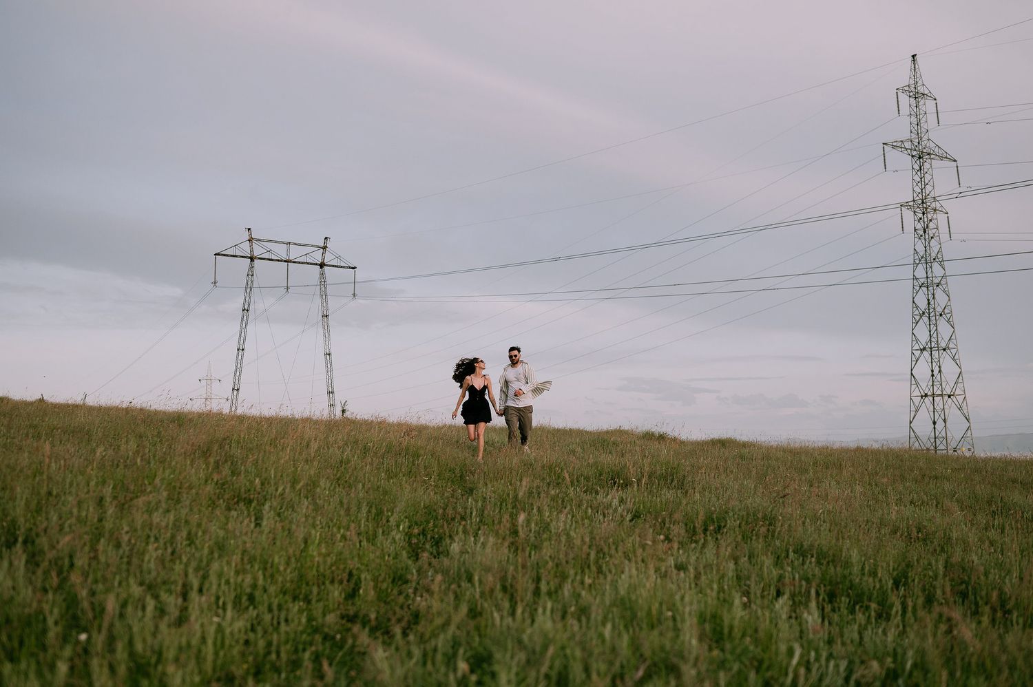 Two silhouettes walk along a grassy hilltop beneath power lines and cloudy skies at dusk.