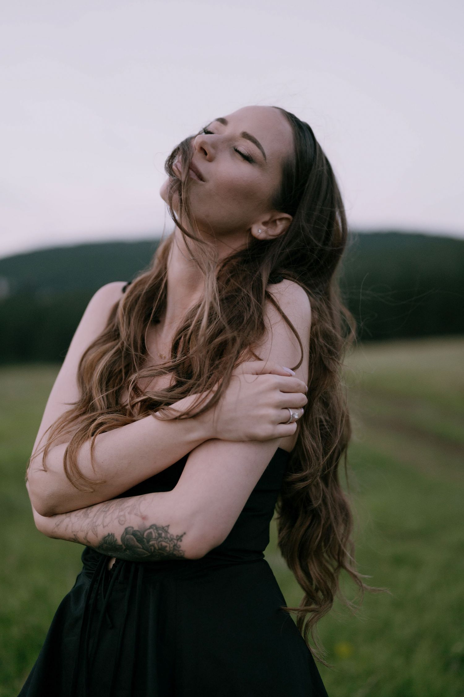 A person in a black dress poses expressively in a field with dramatic body movements and flowing hair.