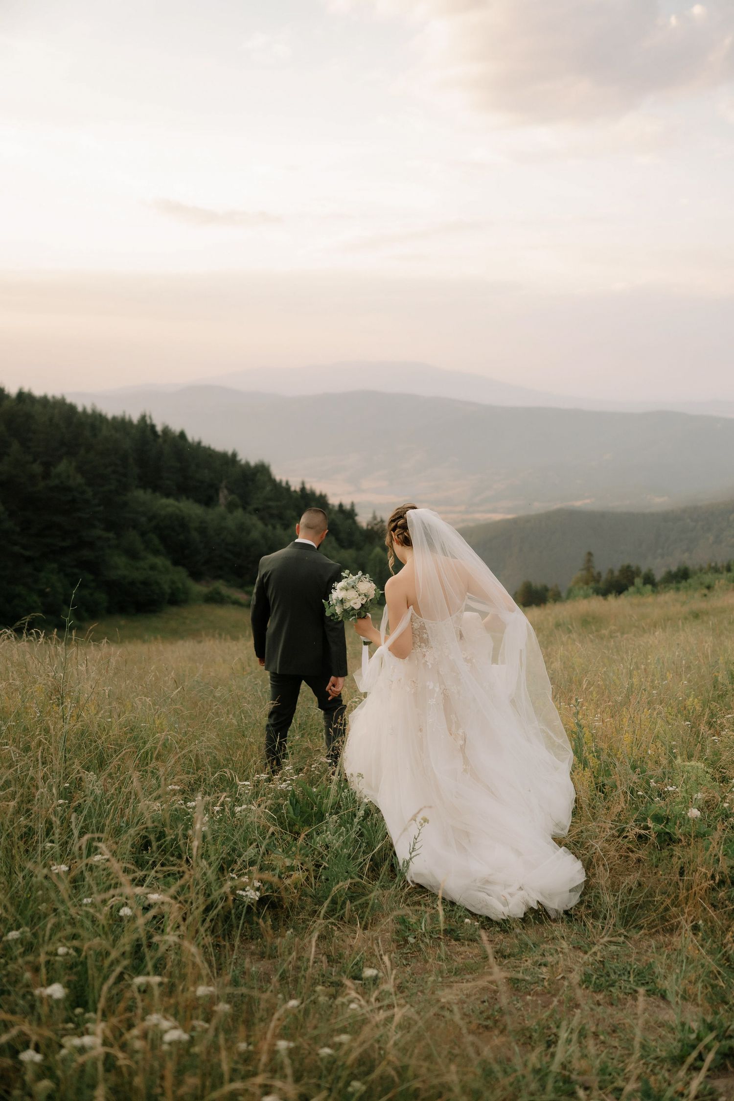 Newlywed couple walks through a misty mountain meadow at sunset in their wedding attire.