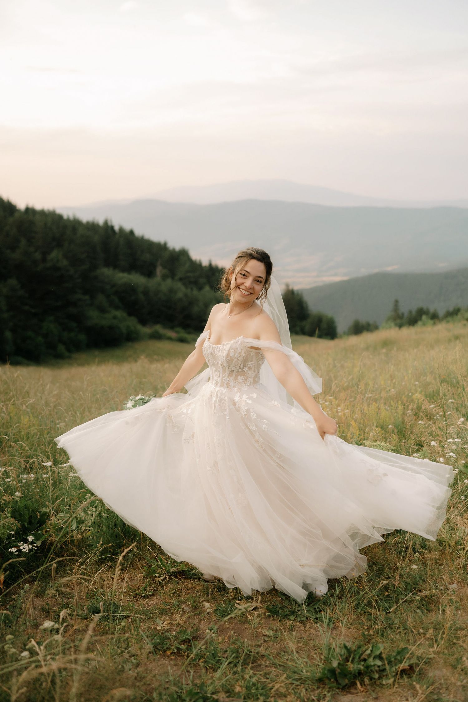 A bride in a white dress spins joyfully in a mountain meadow with wildflowers and rolling hills in the background.
