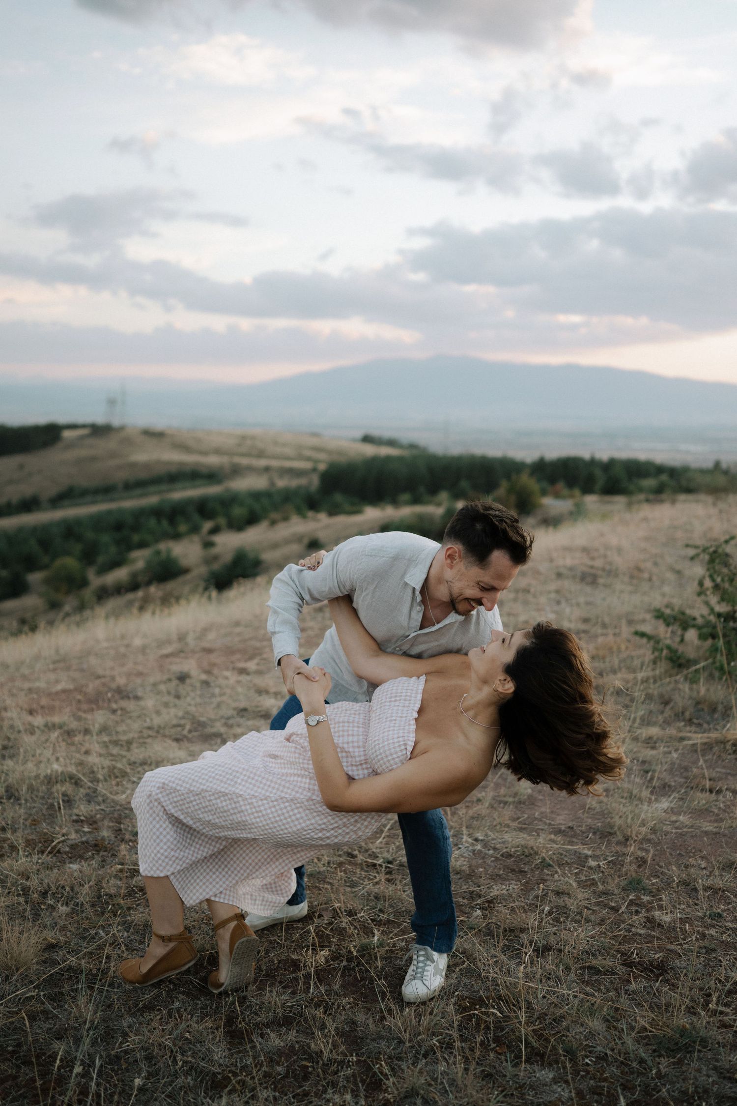 A couple shares a romantic dip-style dance pose against a scenic hillside backdrop at sunset.