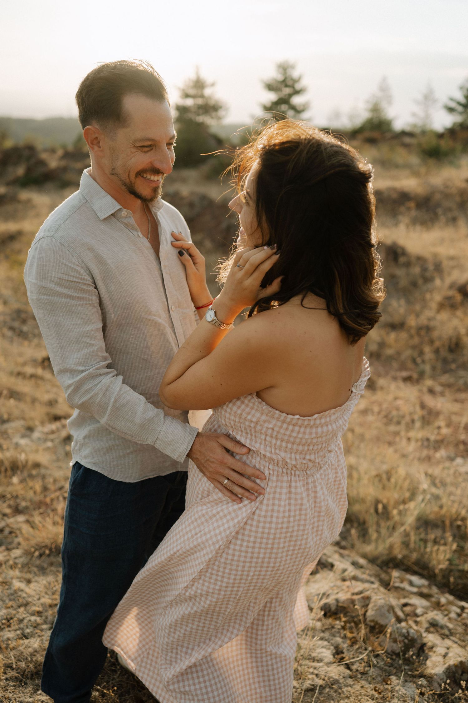 A romantic couple embraces during golden hour in a scenic outdoor setting with mountains and trees in the background.