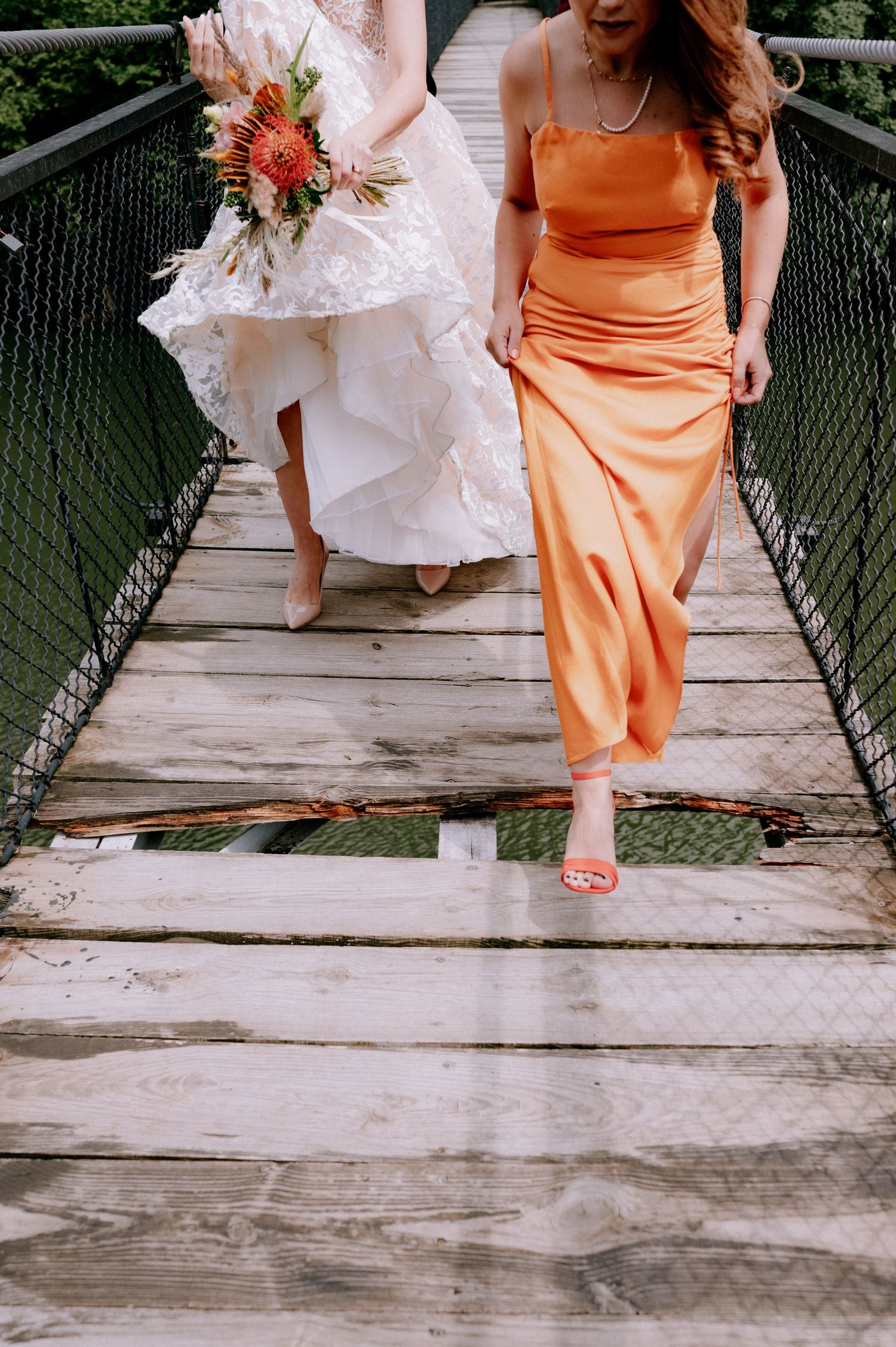 Two people in wedding attire walk across a wooden bridge, one in white and one in orange dresses.