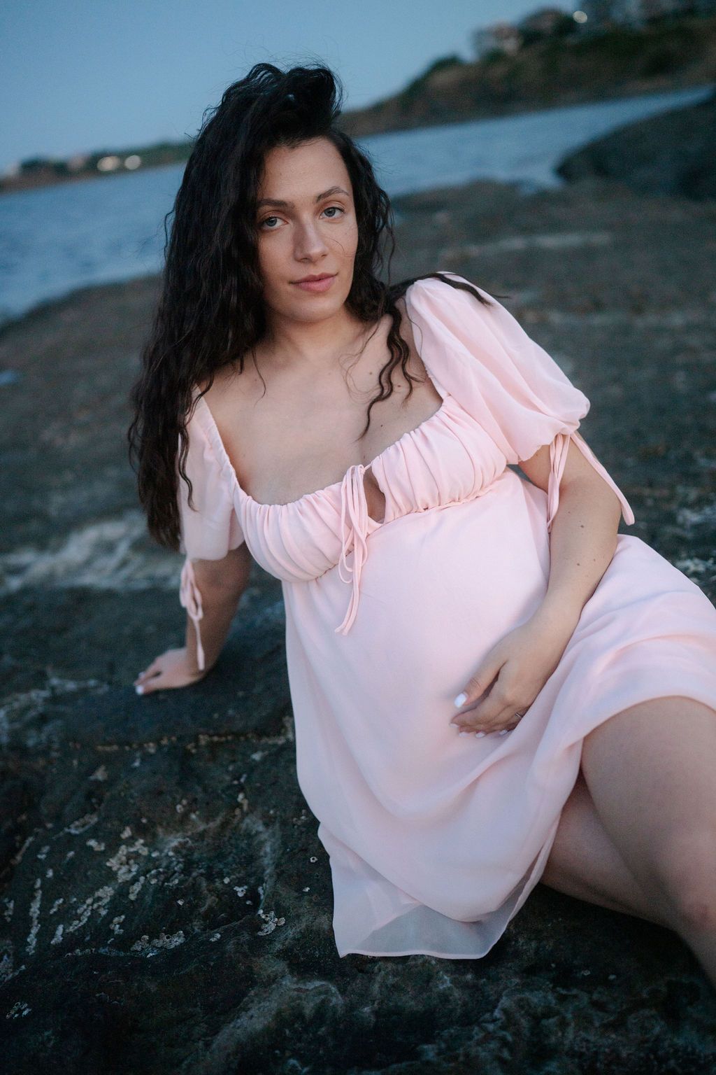 Woman in pink blouse poses on rocky beach during sunset photoshoot.