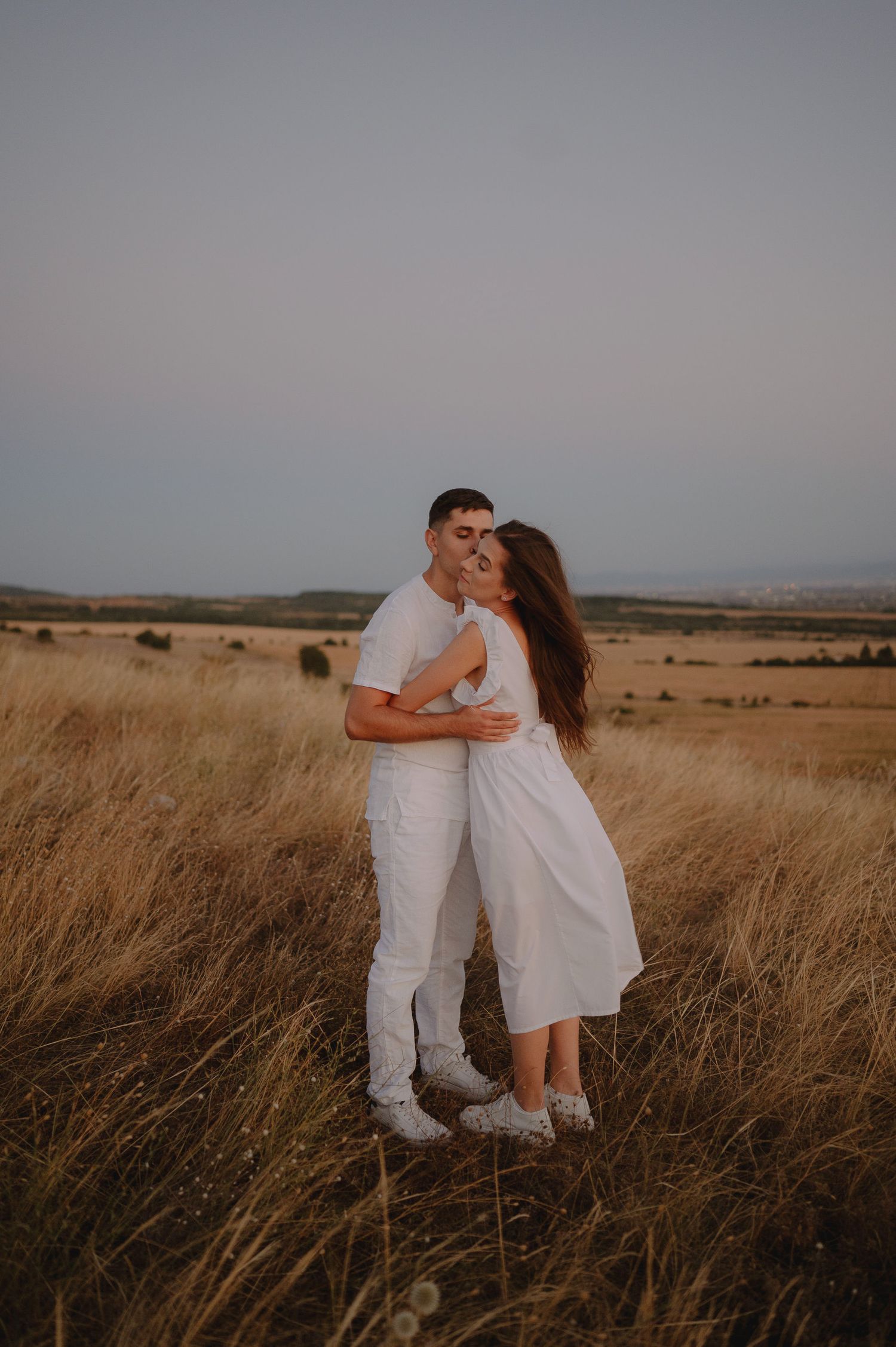 Couple in white outfits embracing in a golden grass field at sunset, creating a romantic silhouette against the horizon.