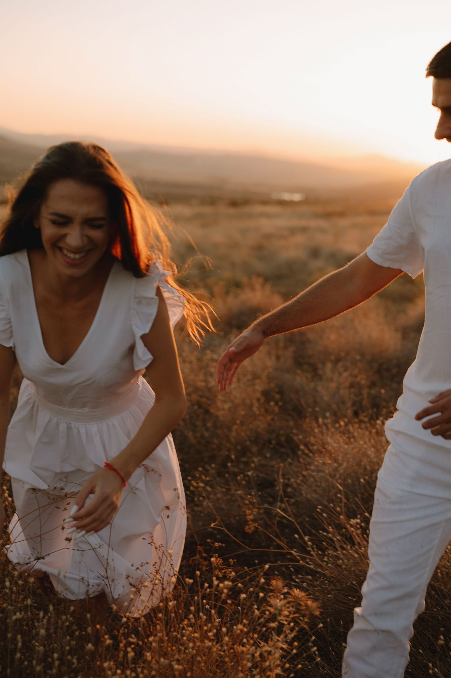 A couple in white outfits walks through a golden field during sunset creating a romantic silhouette.
