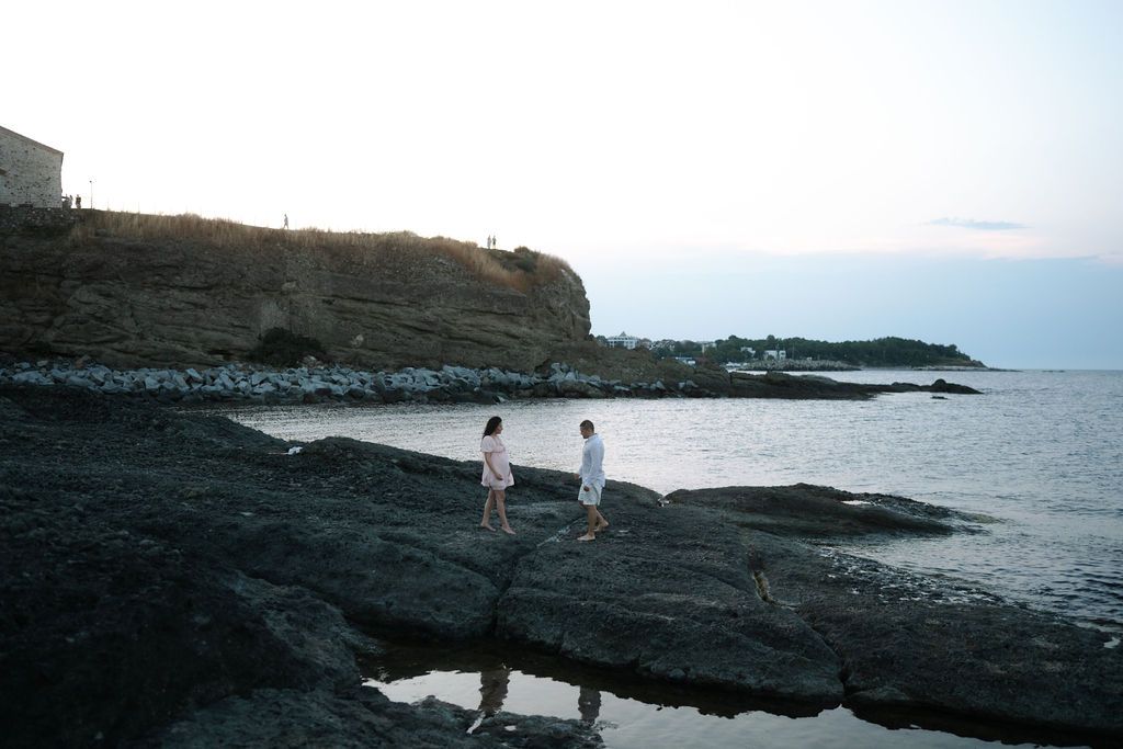Two people stand on dark volcanic rocks overlooking a serene ocean view at sunset.