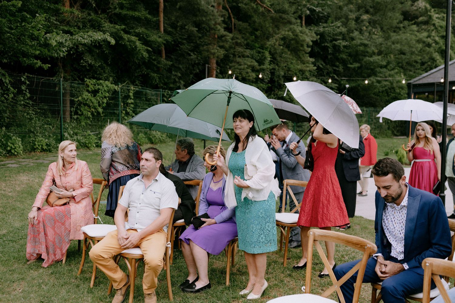 Wedding guests hold white parasols while gathering outdoors in a lush green garden setting during a summer celebration.