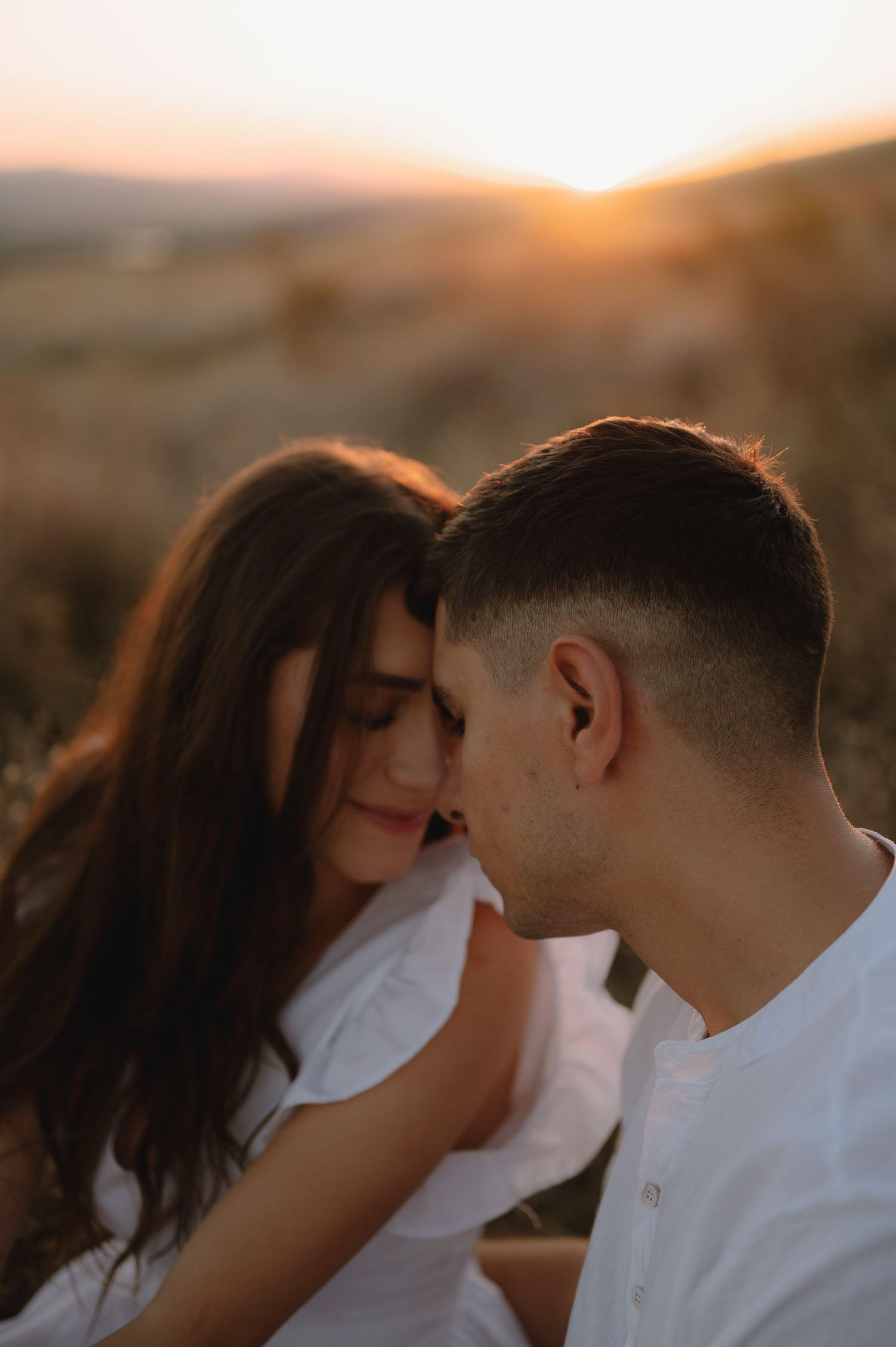A couple in white attire share a romantic moment at sunset with warm golden lighting in the background.