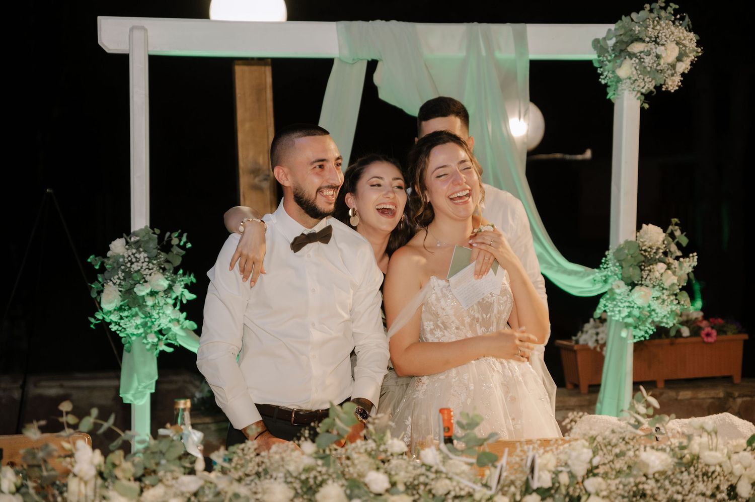Wedding party laughs together under a decorated white arch with green lighting and floral arrangements at night.