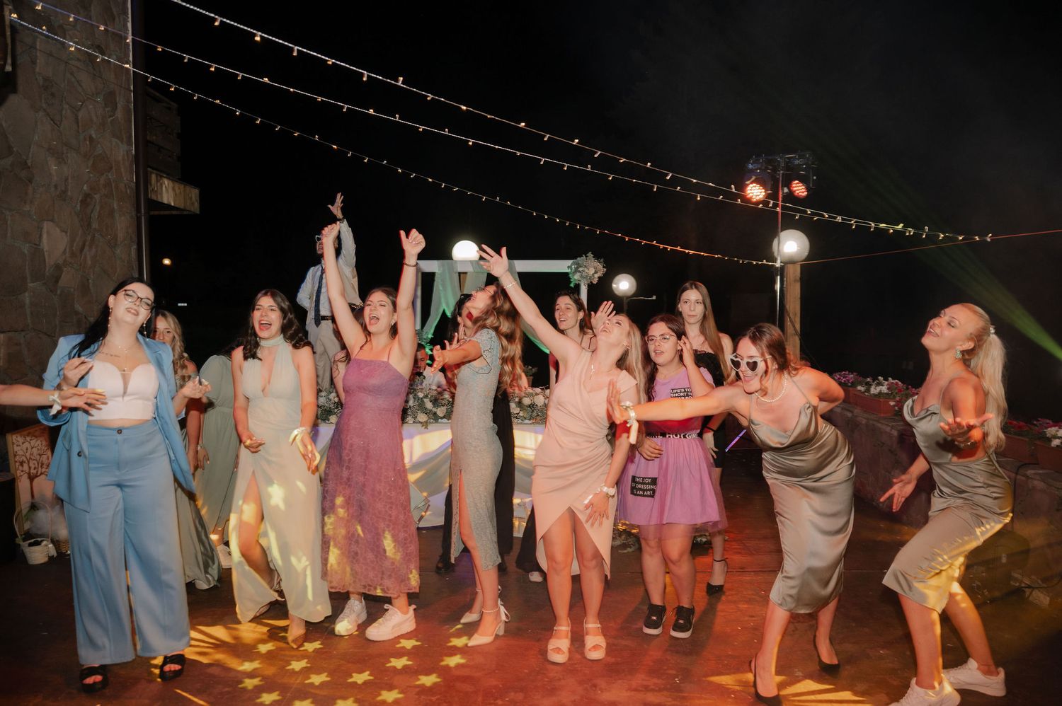 A lively nighttime outdoor party with string lights and people dancing together on a wooden deck.