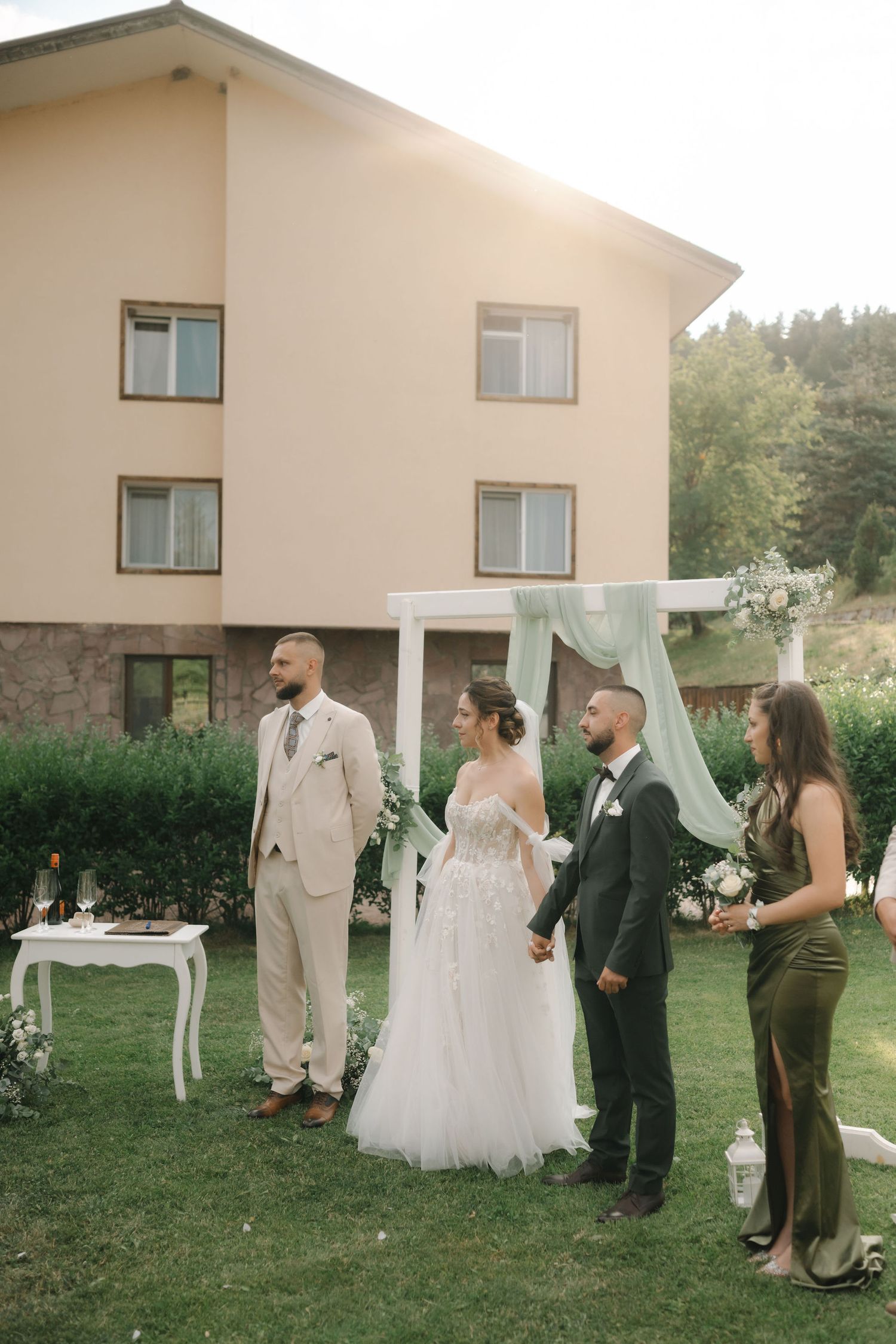Wedding party in formal attire stands on a lawn outside a white building during an outdoor ceremony.