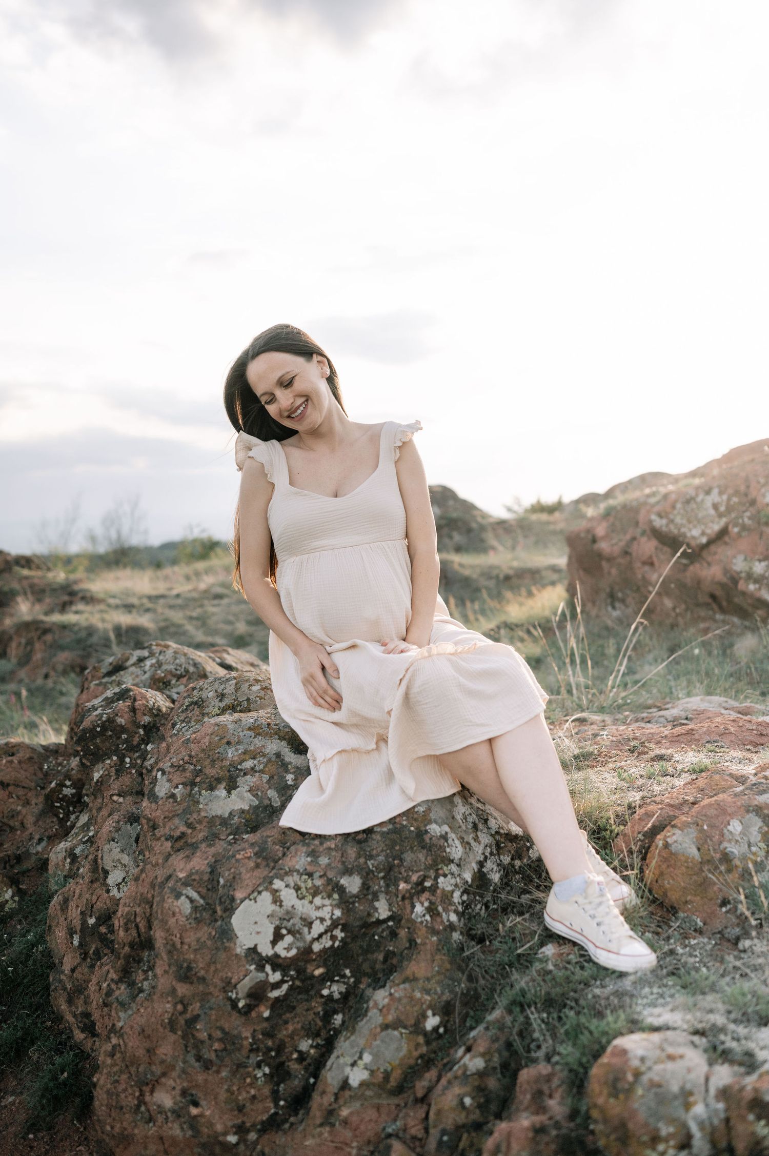 A person in a white dress reclines gracefully on rocky terrain against a cloudy sky in an artistic outdoor photoshoot.
