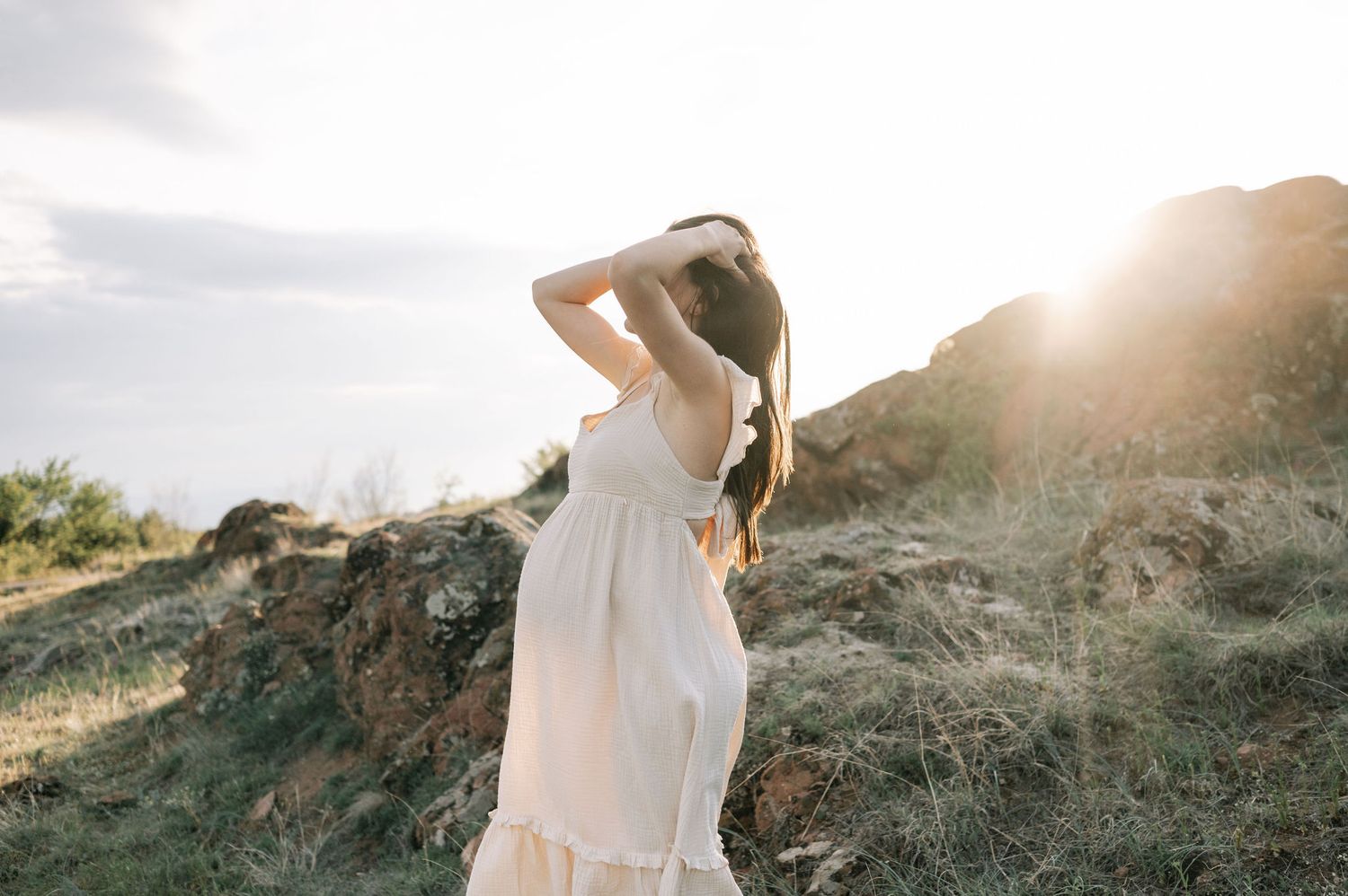 A person in a flowing white dress stands in golden sunlight on a grassy hillside during sunset.