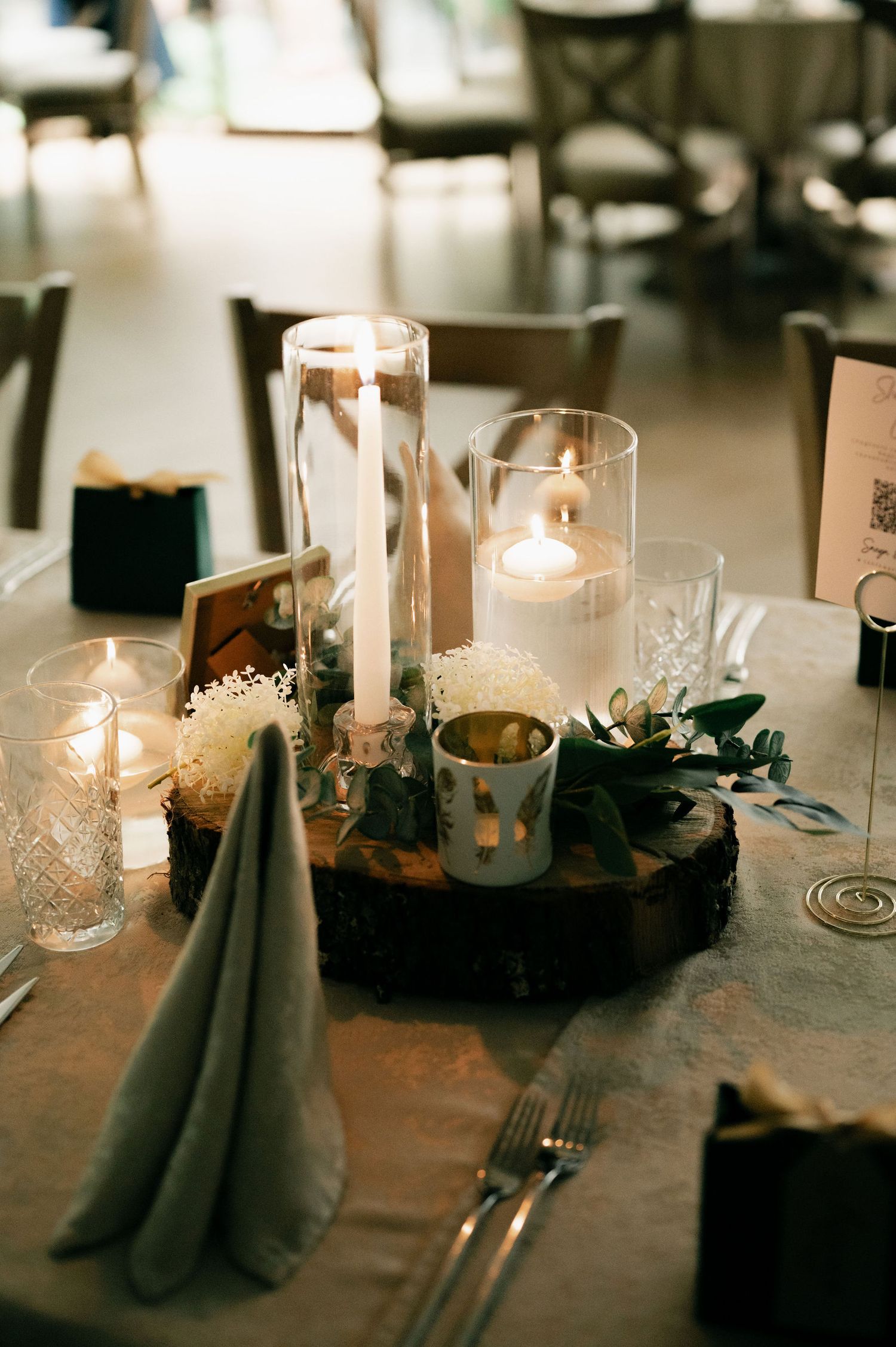 Rustic wedding table centerpiece with glowing candles, greenery and wood slice base on gray linen tablecloth.