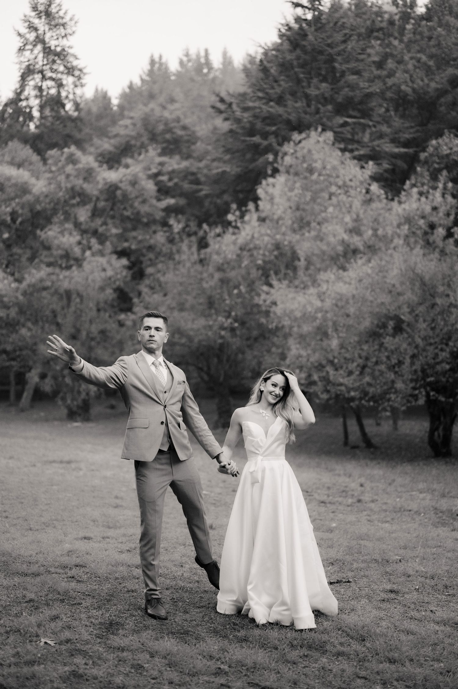 A couple dances joyfully in a meadow surrounded by evergreen trees in black and white photography.