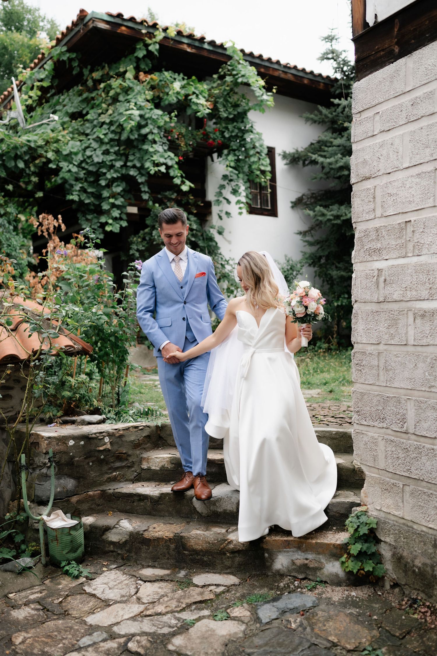 A couple in wedding attire descends stone steps at a rustic venue with climbing vines and greenery.