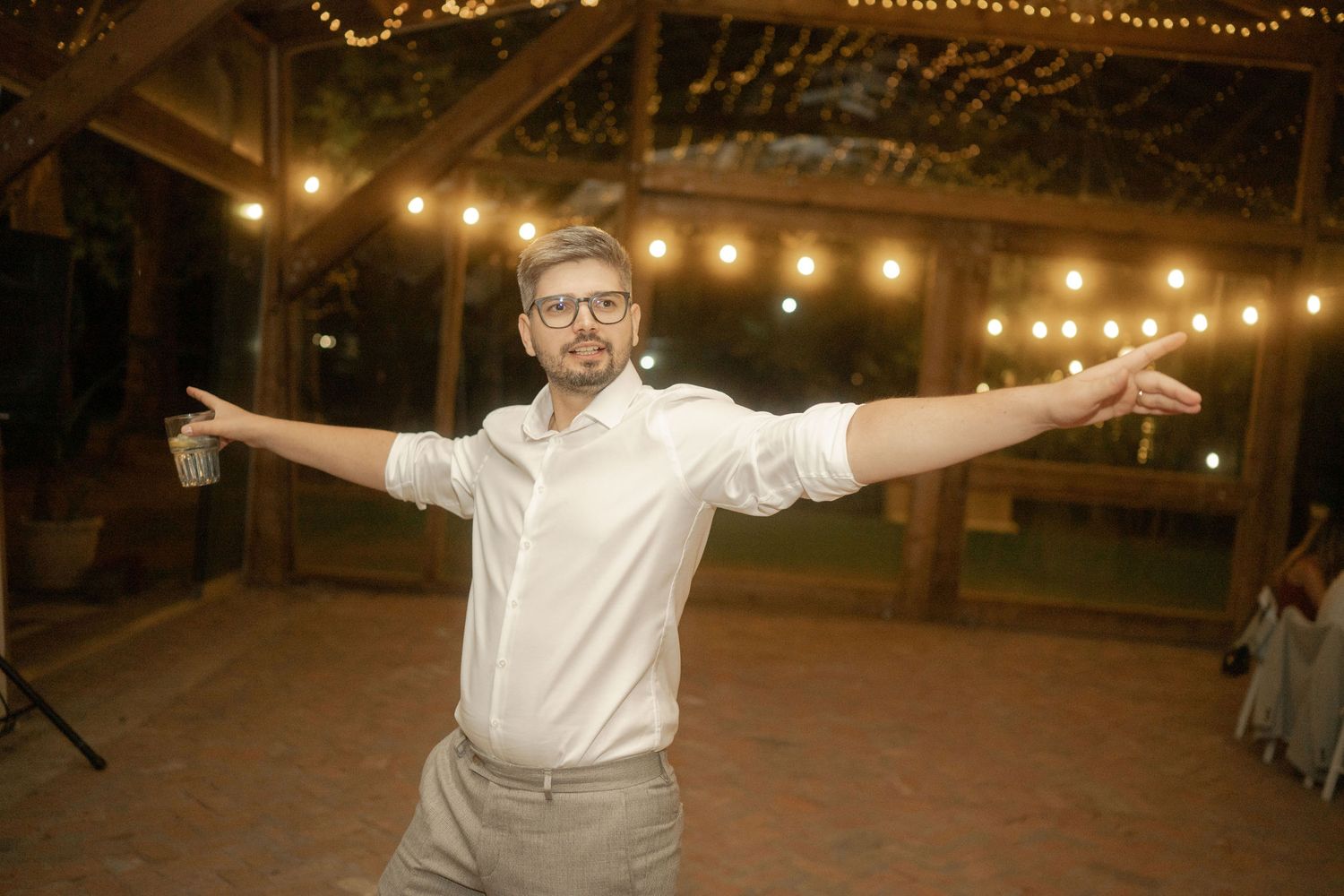 A person dances joyfully with arms spread wide under string lights at an indoor venue.