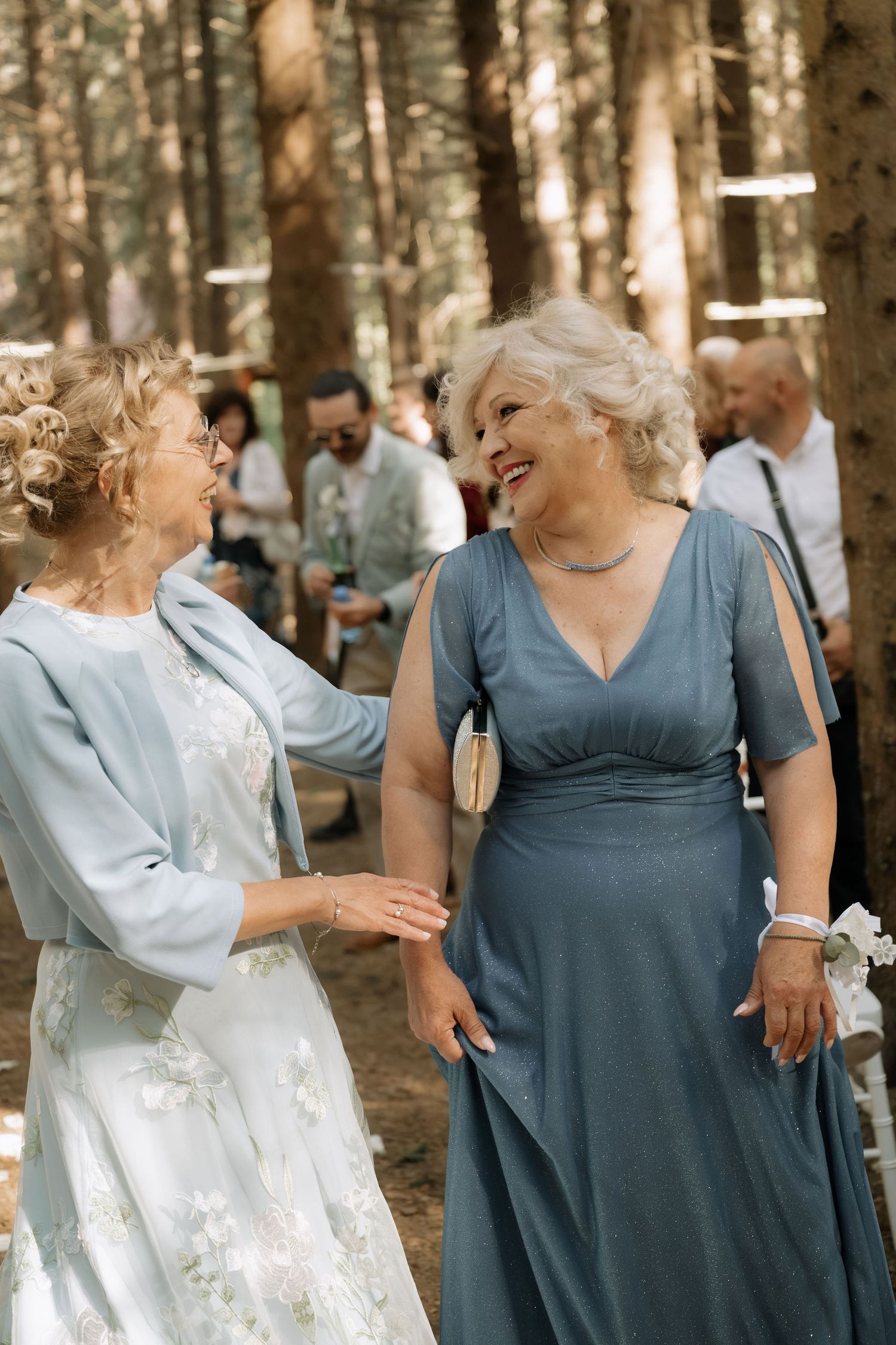 Two elegant older women in blue dresses laughing together at an outdoor woodland wedding celebration.