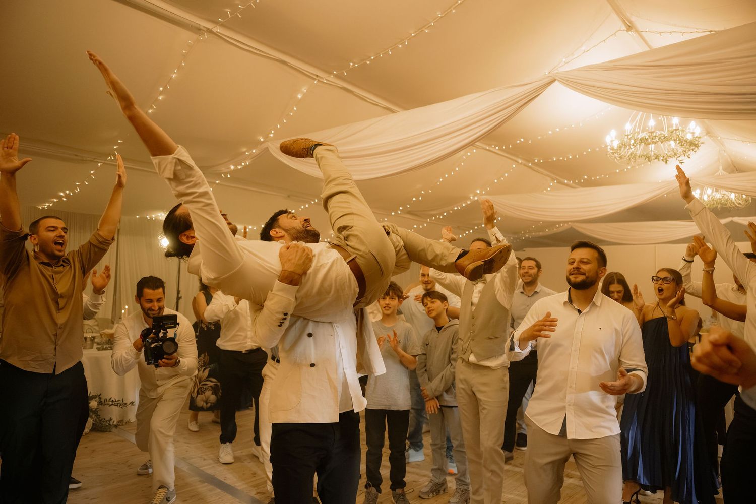 Guests celebrate at a wedding reception, lifting someone overhead on the dance floor under draped fabric lights.