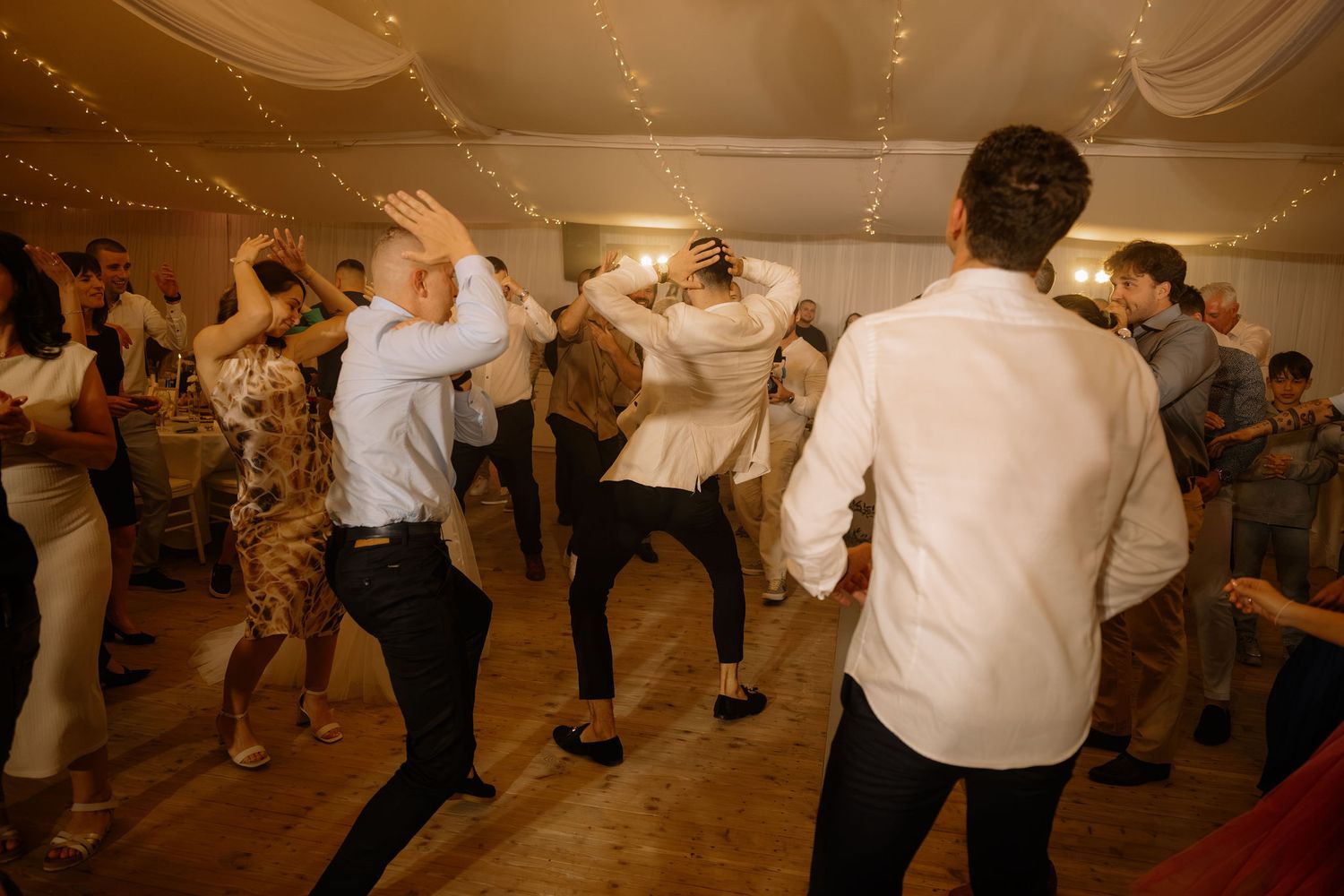 Guests dancing energetically at a wedding reception in a decorated tent venue with string lights.