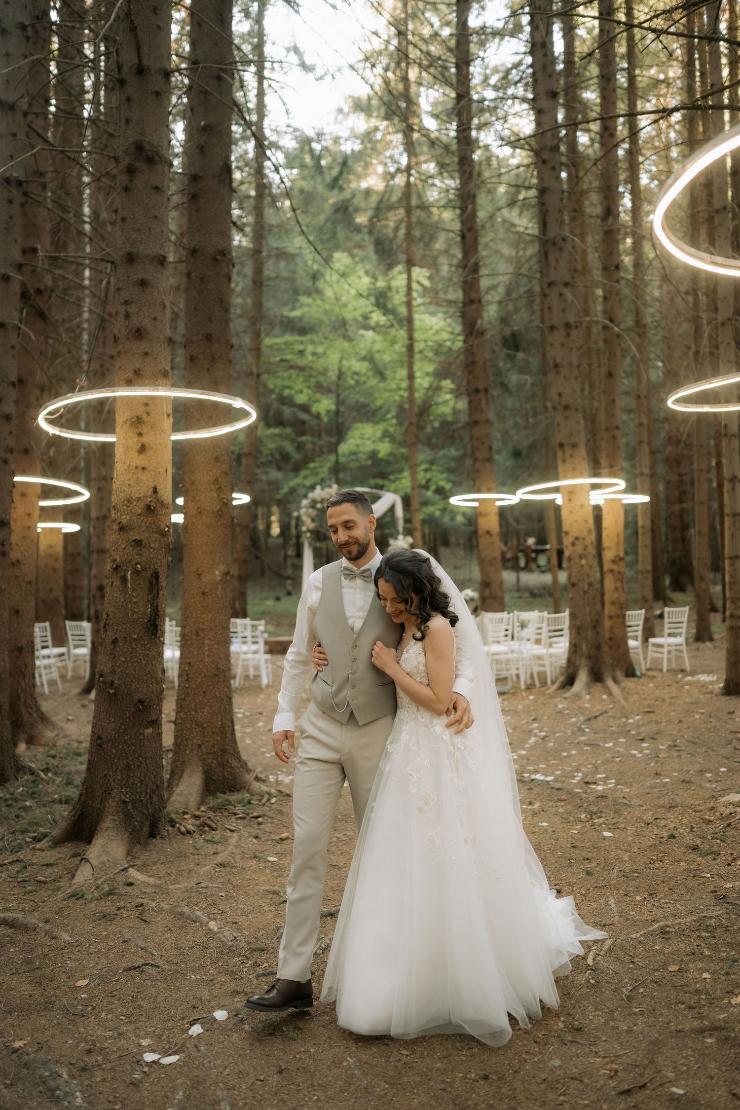 Bride and groom embrace in an enchanting forest wedding venue with glowing circular lights hanging from tall trees.