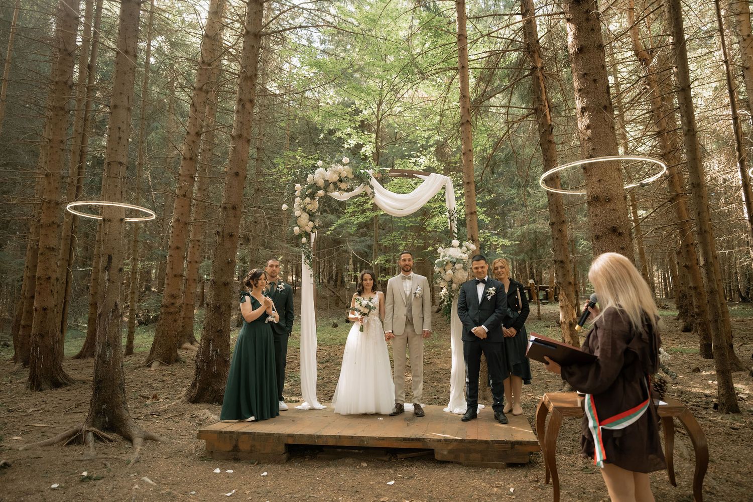 Outdoor forest wedding ceremony on wooden platform with floral arch, wedding party in green and navy, surrounded by tall pine trees.