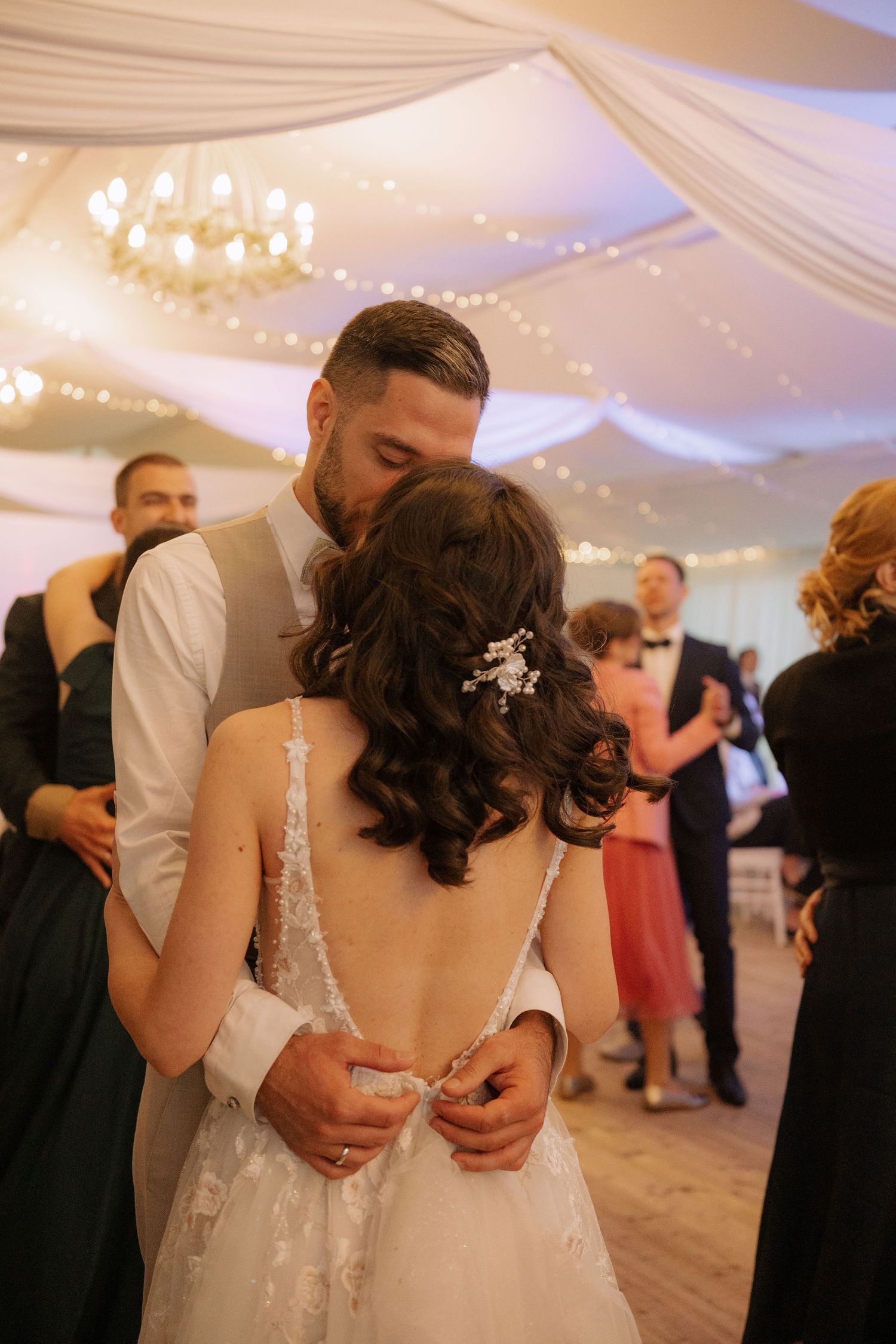 Bride and groom share first dance kiss at elegant wedding reception with fairy lights and chandelier.