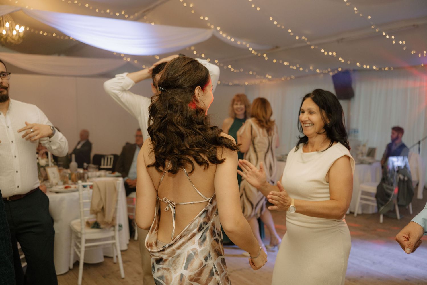 Guests dancing joyfully at a wedding reception under warm string lights in an elegant white tent venue.