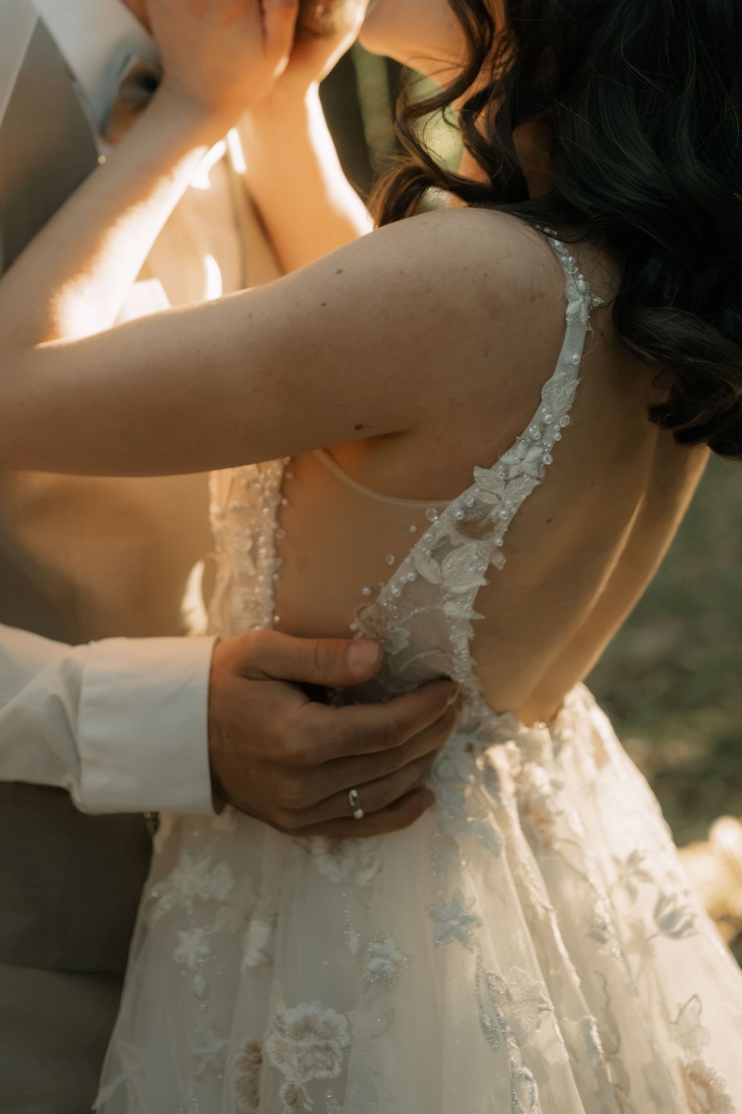 Groom fastening buttons on brides lace wedding dress from behind in warm golden sunlight.