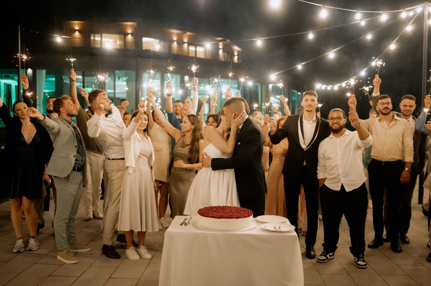 Wedding guests celebrate with raised hands around a cake table under romantic string lights at an outdoor evening reception.