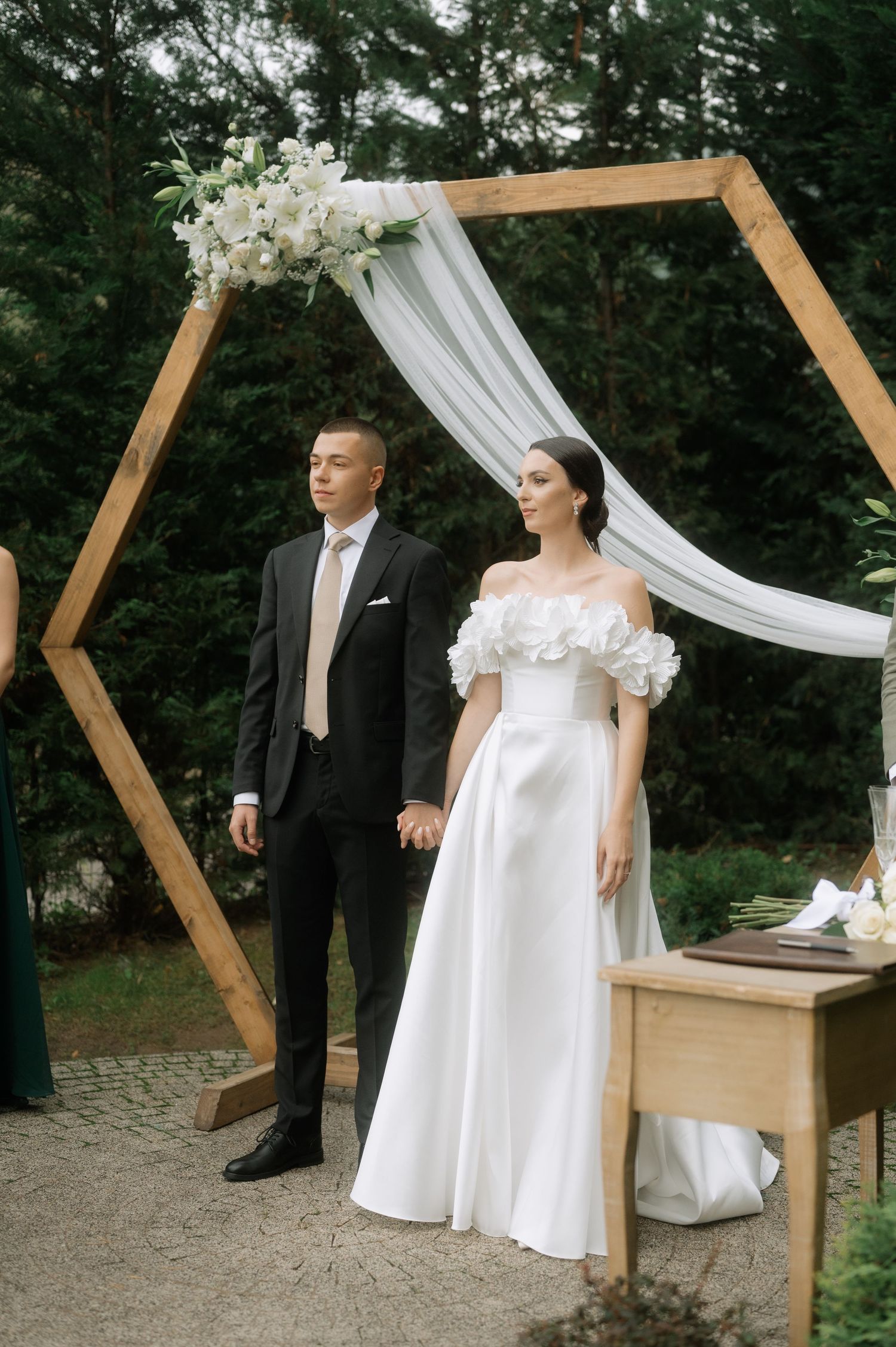 Couple stands beneath a wooden hexagonal wedding arch draped with white fabric during an outdoor forest ceremony.