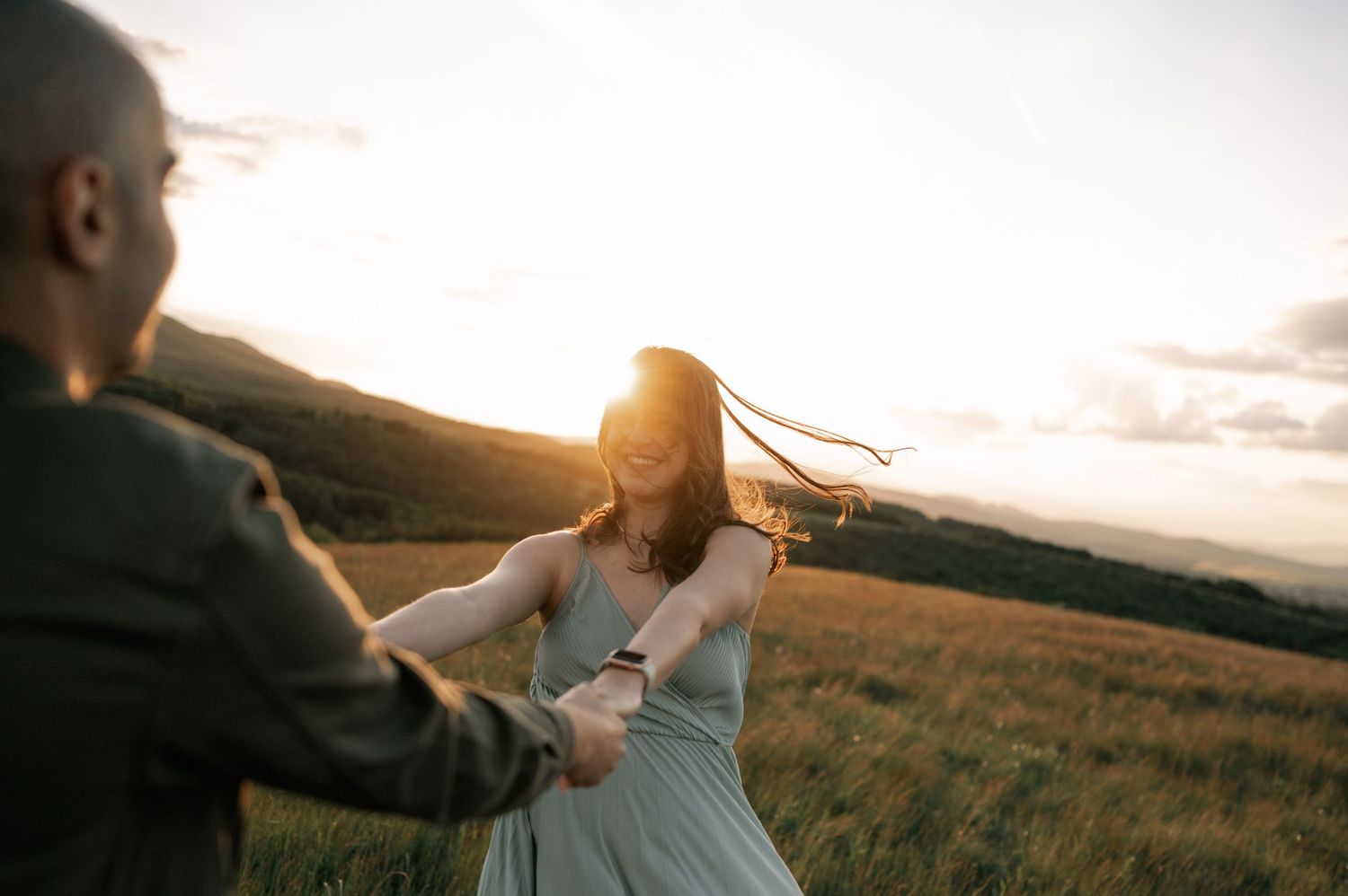 Couple dancing in golden sunset field, woman in blue dress with flowing hair, romantic outdoor engagement photo.