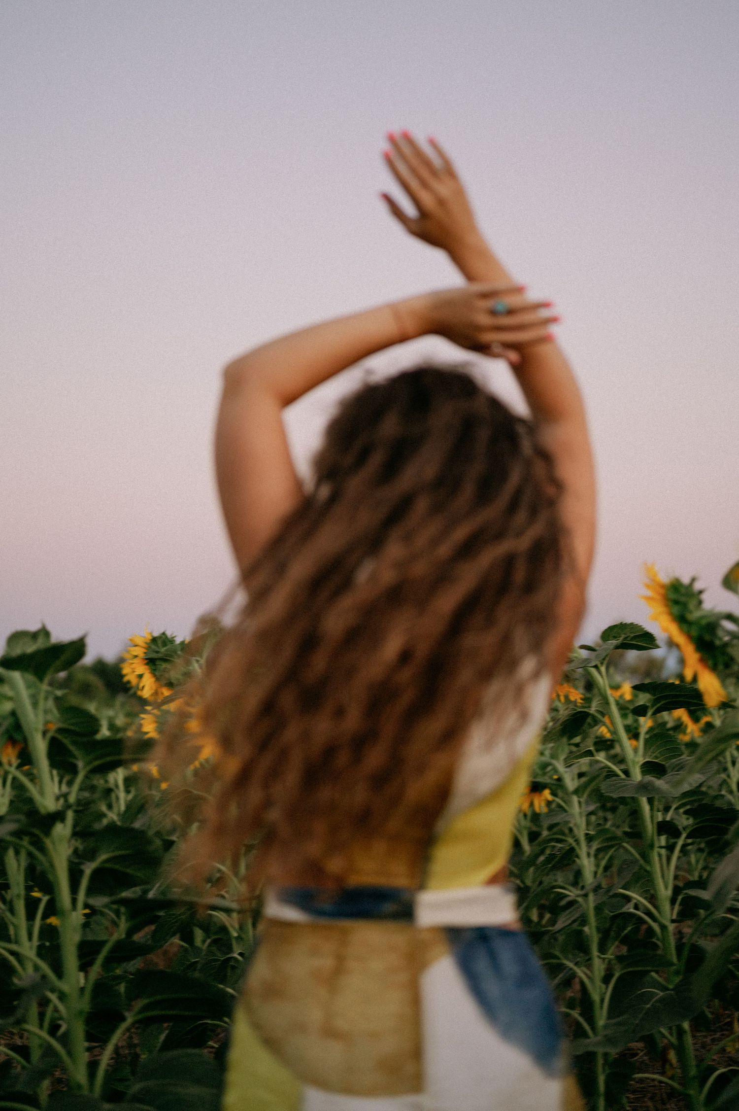 Woman with curly hair raises arms in a sunflower field at dusk, wearing a yellow top and denim.