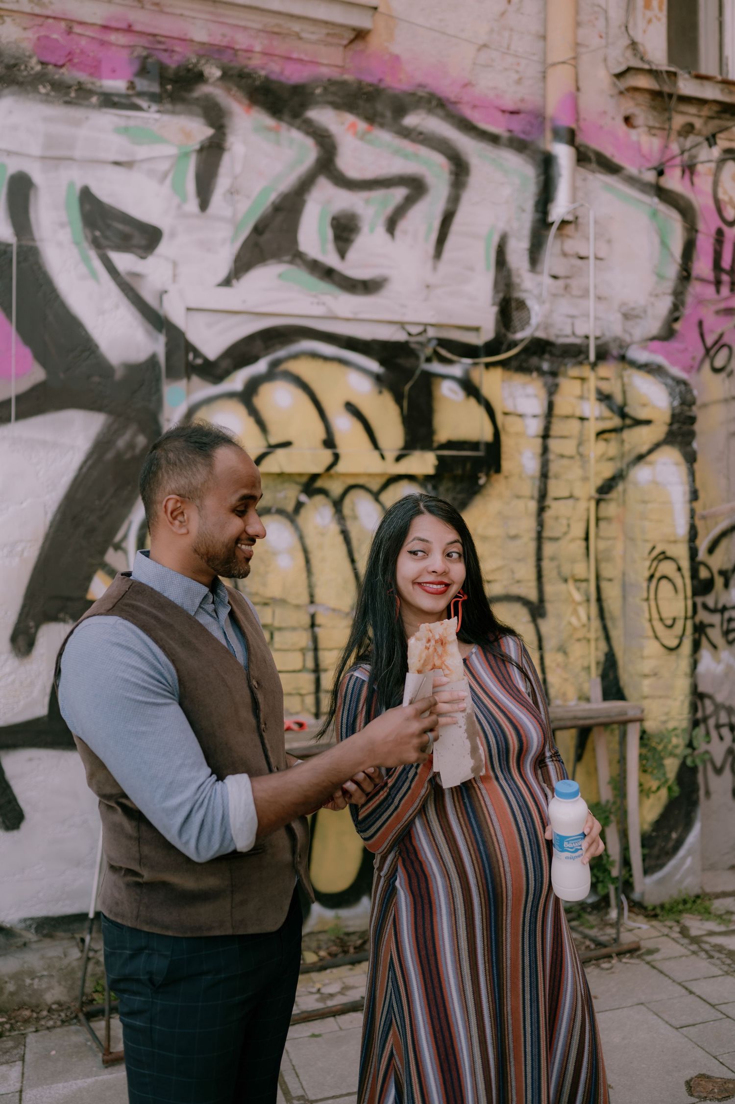 Pregnant woman in striped dress shares food with man in vest against colorful graffiti wall outdoors.