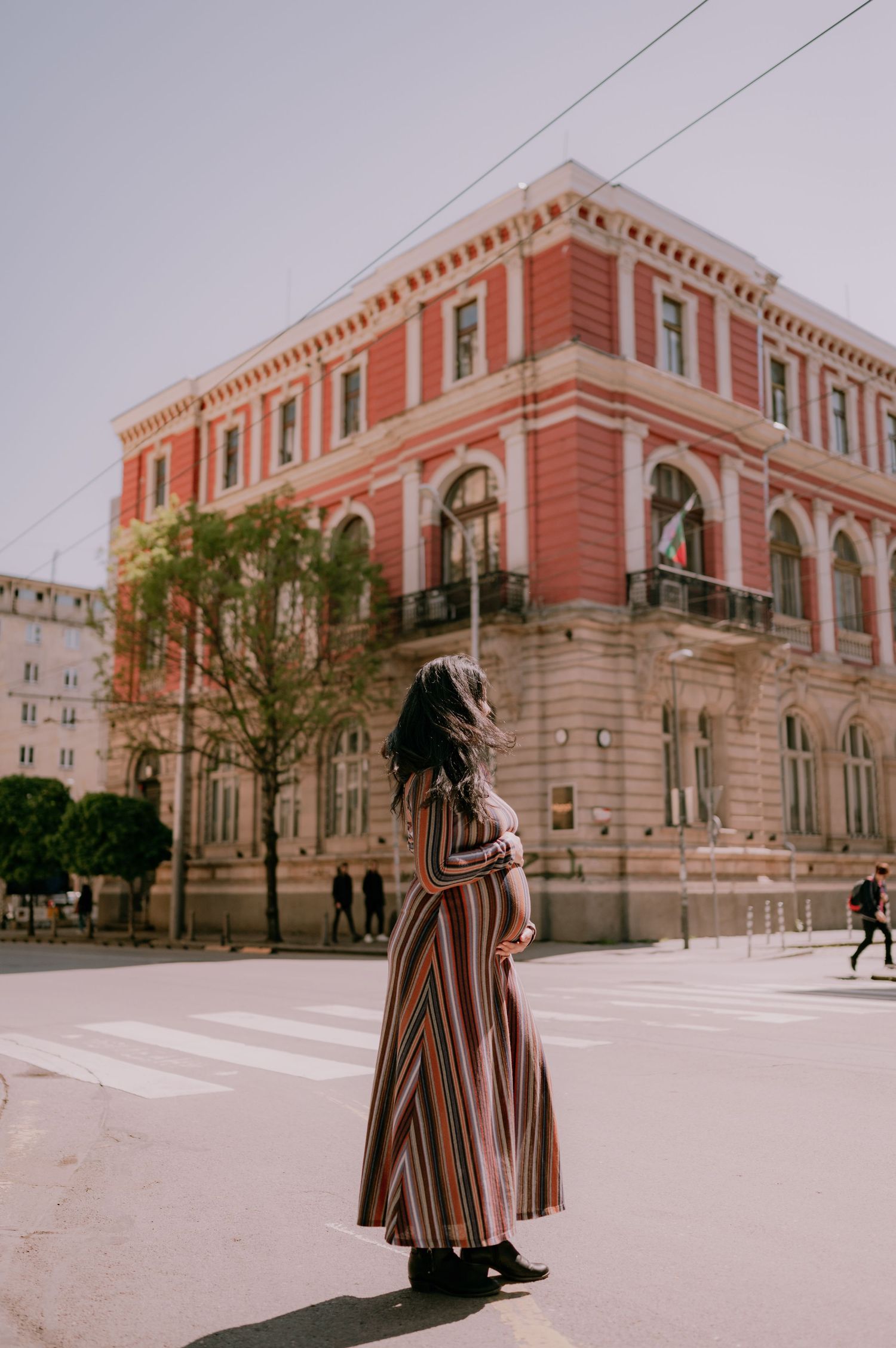 Woman in striped maxi dress standing on urban crosswalk with red historic building in background.