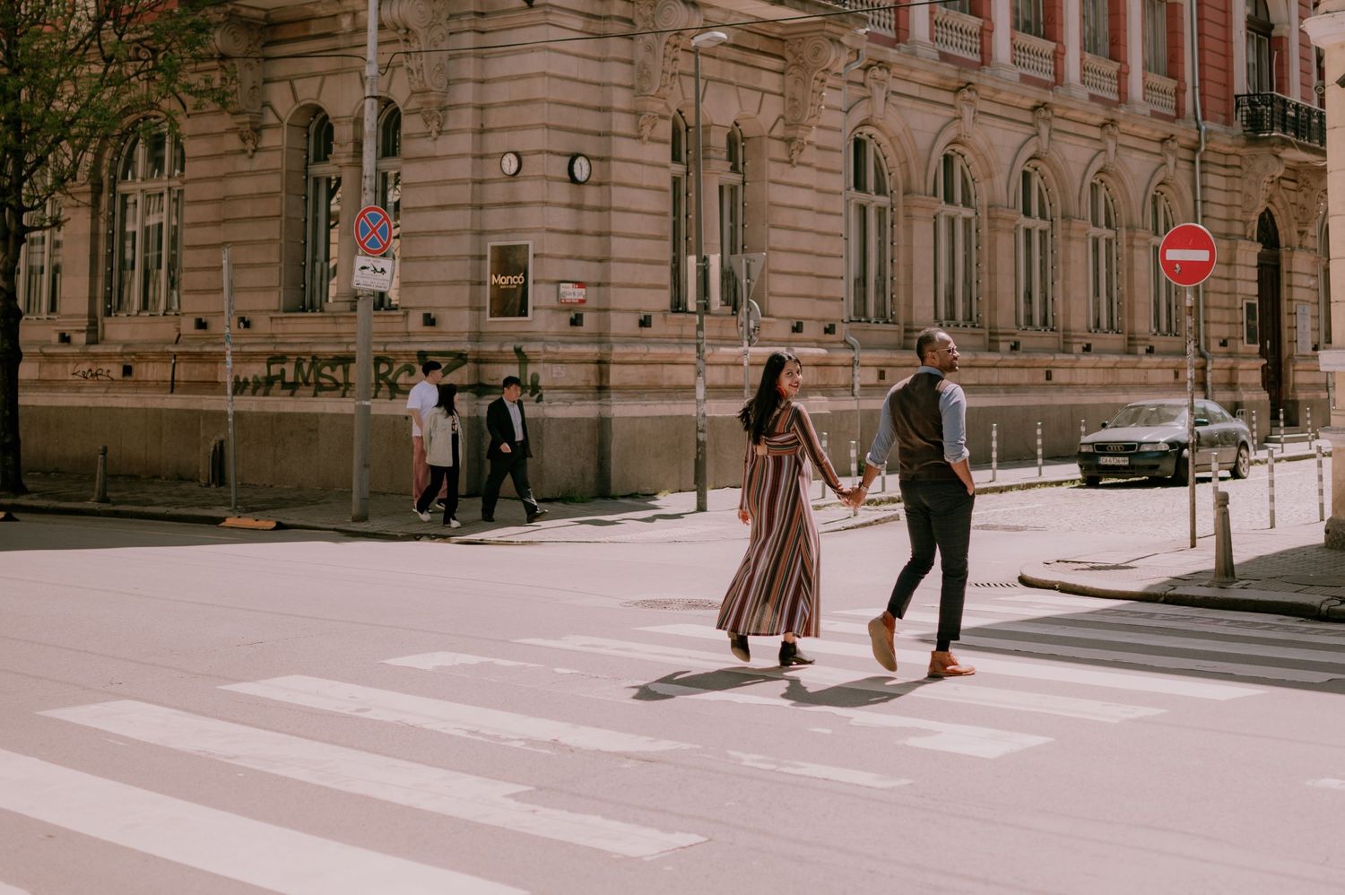 Couple holding hands crossing a zebra crossing in front of a historic building on a sunny urban street.