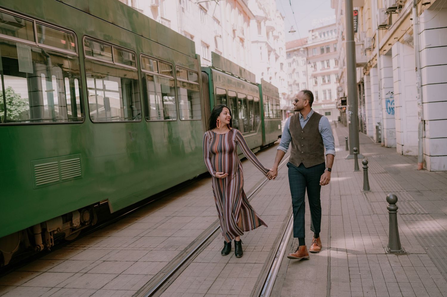 Couple holding hands walking beside a green tram on a city street, dressed in stylish outfits on a sunny day.