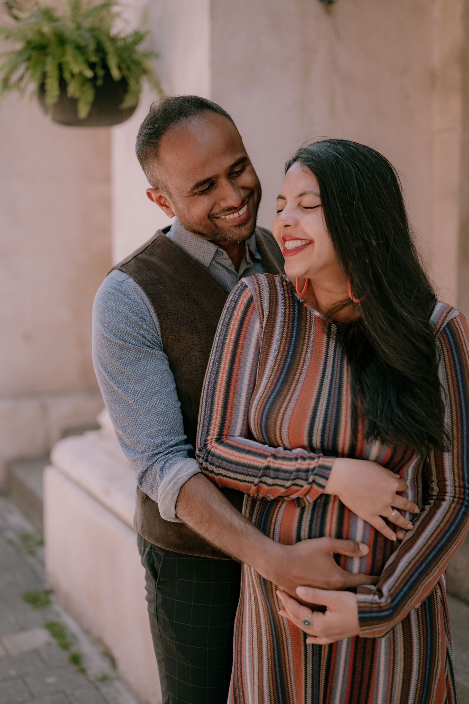 Smiling couple embracing outdoors, pregnant woman in striped dress with eyes closed, man in vest hugging her baby bump.